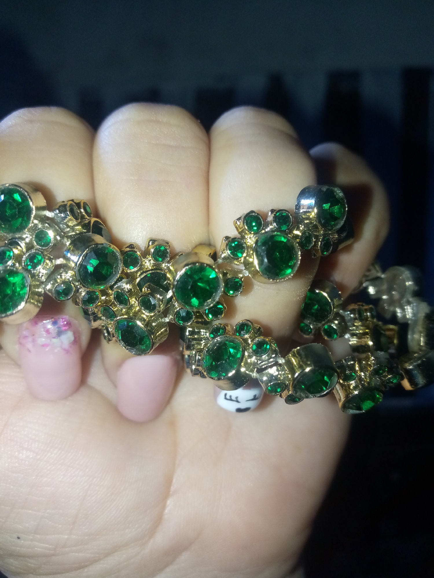 A close-up shot depicts a hand adorned with multiple gold-toned rings featuring green gemstones. The rings are clustered on several fingers. The nails are manicured. One nail has a floral design while another has a cartoon face. The background is dark and indistinct. The location is unspecified.