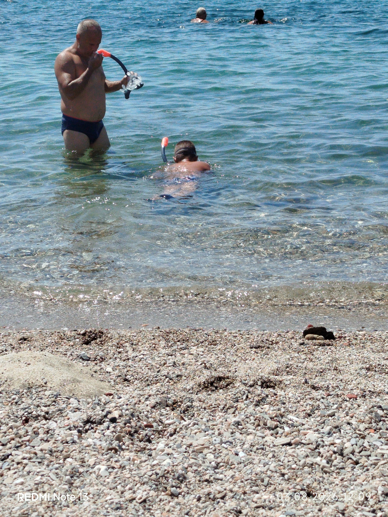 The image depicts a sunny day at a beach with clear, blue water. In the foreground, a pebbly shoreline is visible. In the middle ground, a man in dark swimming trunks is standing in the shallow water, holding a snorkel and mask. He appears to be preparing to snorkel. To his right, another person, likely a child, is partially submerged in the water, also with a snorkel. Further in the background, two other individuals are swimming in the sea. The overall scene suggests recreational activity at a beach, likely in a coastal area. The location is identified as Sithonia Municipal Unit, Greece.