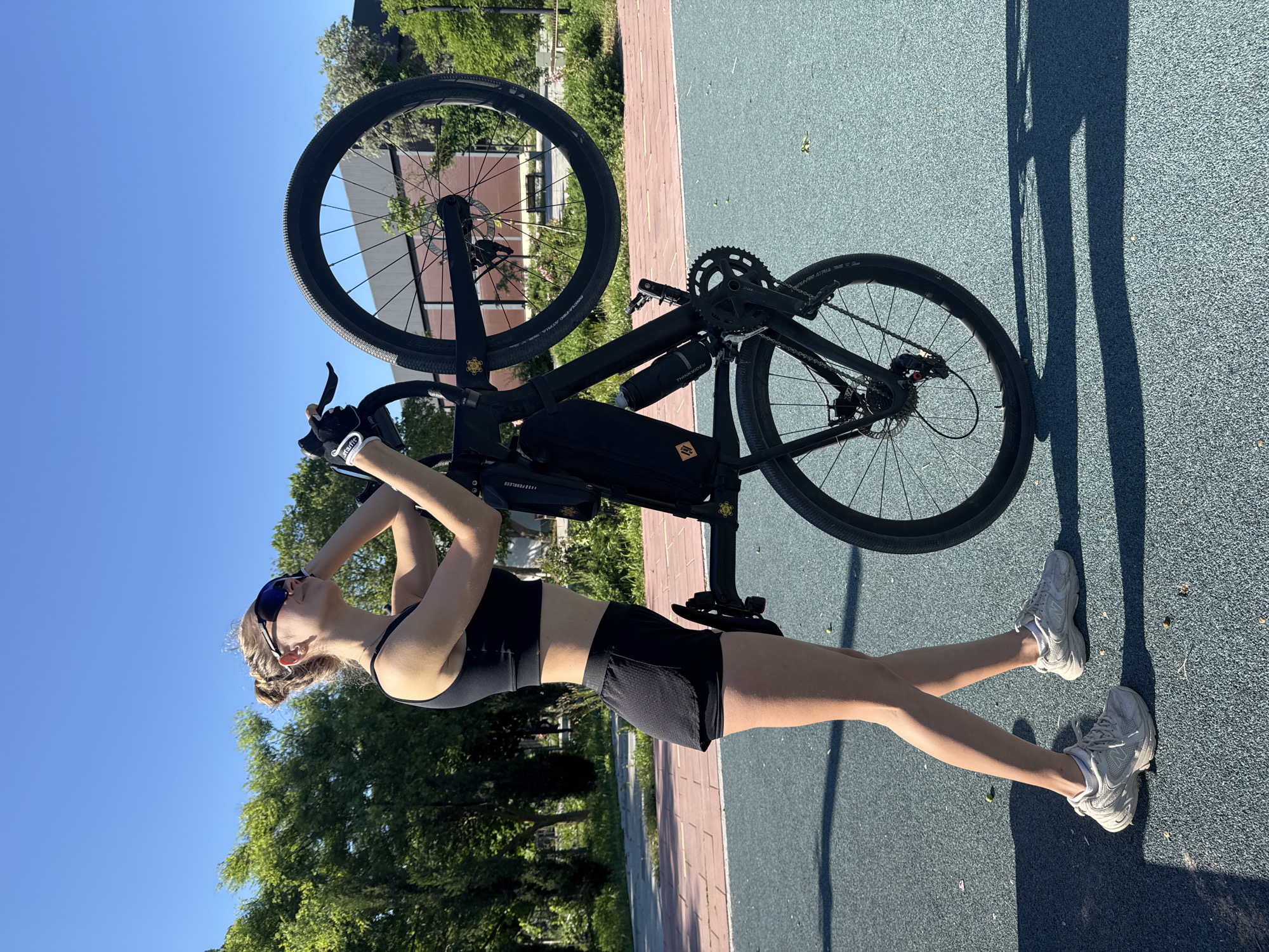 A young woman is holding a black gravel bicycle above her head outdoors. She is wearing a black sports bra, black shorts, white running shoes, and blue-tinted sunglasses. Her hair is pulled back. She is standing on a surface that is part blue and part red, possibly a running track or a recreational area. In the background, there are green trees and a building. The image appears to be taken in bright sunlight. Location: Мысовское сельское поселение, Ukraine.
