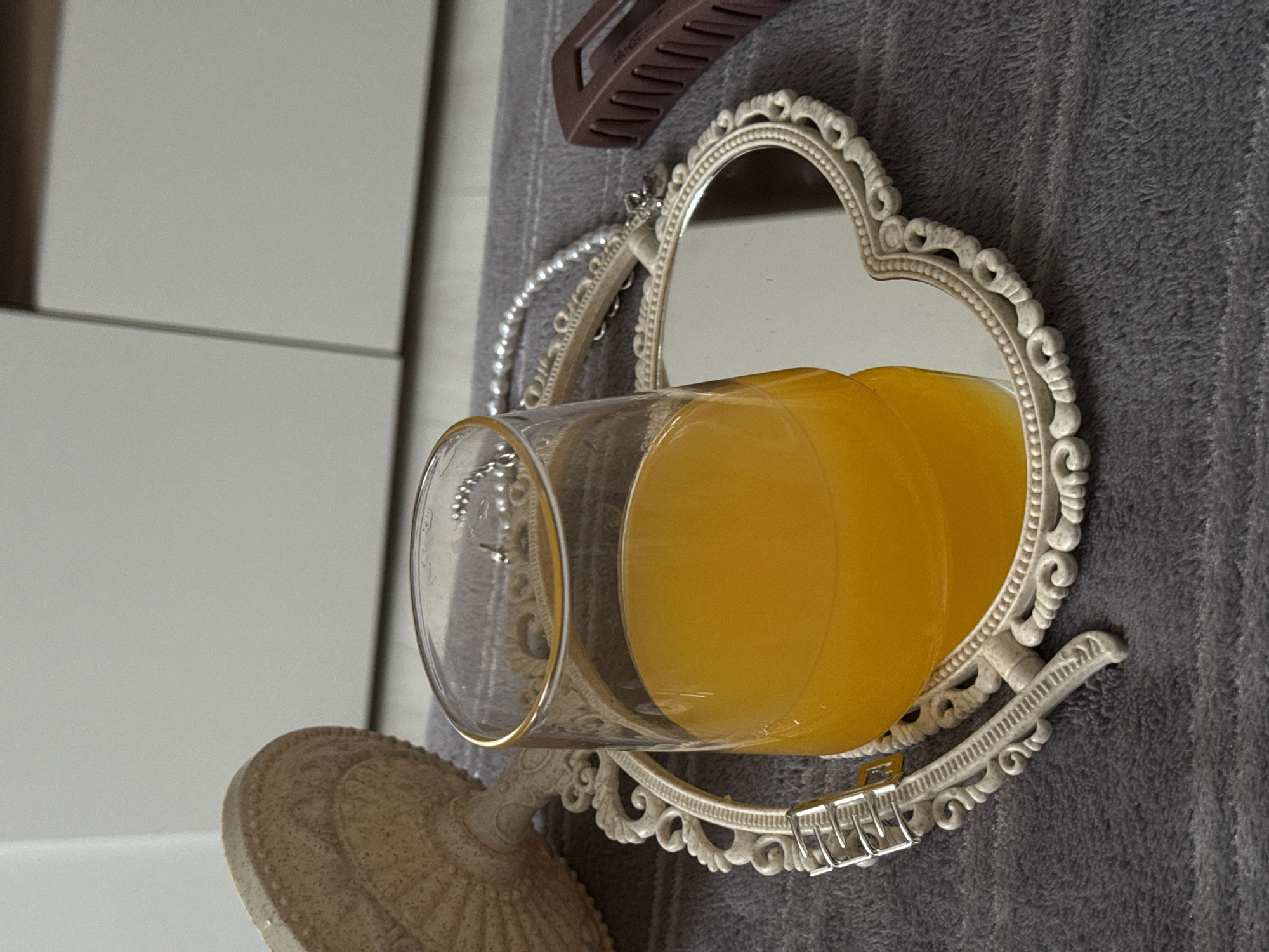 A glass of yellow liquid rests on a heart-shaped decorative mirror that sits on a gray, textured surface. A brown hair clip partially obscures the top of the mirror. A white surface is visible in the background.