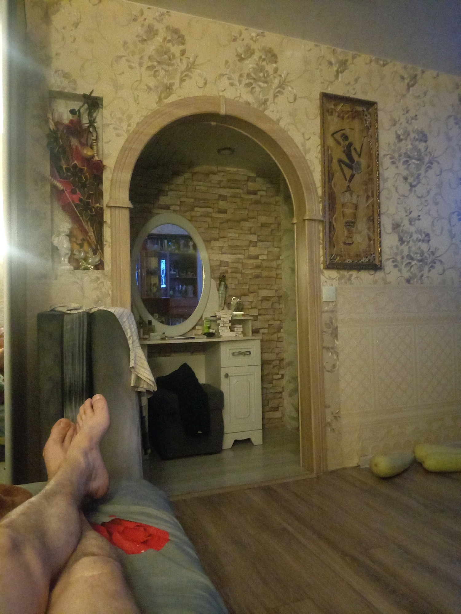 The image depicts an interior scene, possibly a living room, featuring a person's legs and feet in the foreground, resting on what appears to be a sofa or chair. The person is wearing blue pants and a red piece of fabric is visible on the pants. The background reveals an arched doorway leading to another room with a brick wall and a vanity with a large oval mirror. The walls of the main room are adorned with floral wallpaper and framed artwork. Two light-colored, elongated objects, possibly gourds or large fruits, are placed on the wooden floor near the wall. The lighting suggests it is evening or the room is dimly lit. The scene does not contain enough specific information to determine the exact location in the city of Ivanovo, Russia.
