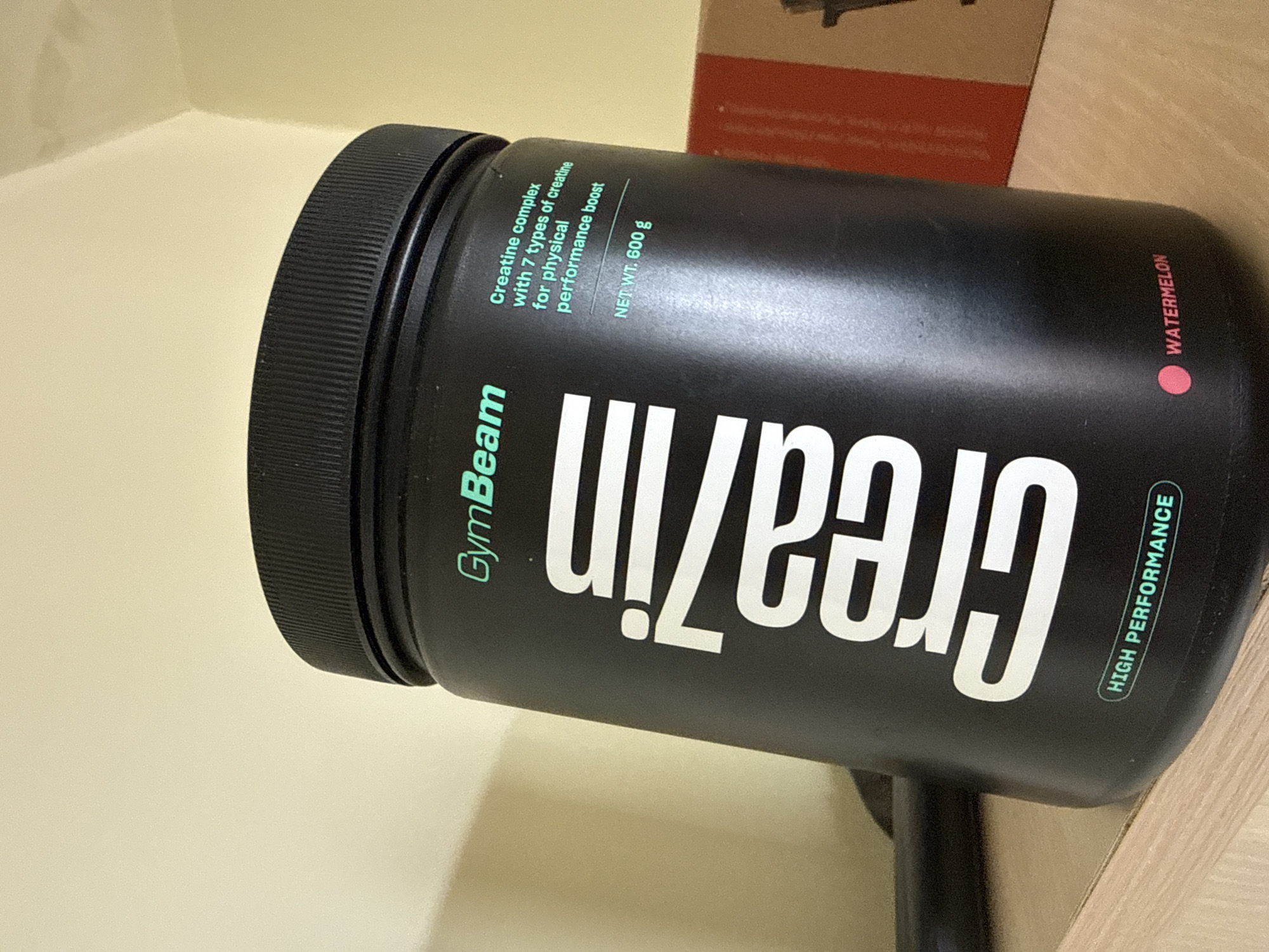 The image displays a black cylindrical container of a supplement, tilted diagonally on a light-colored surface, likely a shelf or table, against a matching light-colored wall. Part of a brown cardboard box is visible in the background.
The primary subject is the black container, featuring a ribbed black lid. The label on the container is predominantly black with text in green and white. The brand name "GymBeam" is visible in green text on the upper left side. The product name "Creazin" is prominently displayed in large white letters across the center.
Smaller green text provides details about the product: "Creatine complex with 7 types of creatine for physical performance boost." Below this, "NET WT. 600 g" indicates the net weight. The flavor, "WATERMELON," is noted in pink text accompanied by a pink dot on the right side of the container. A green outlined box on the lower right states "HIGH PERFORMANCE." The setting is indoors, with no specific activity taking place beyond the product being displayed. No people are visible.