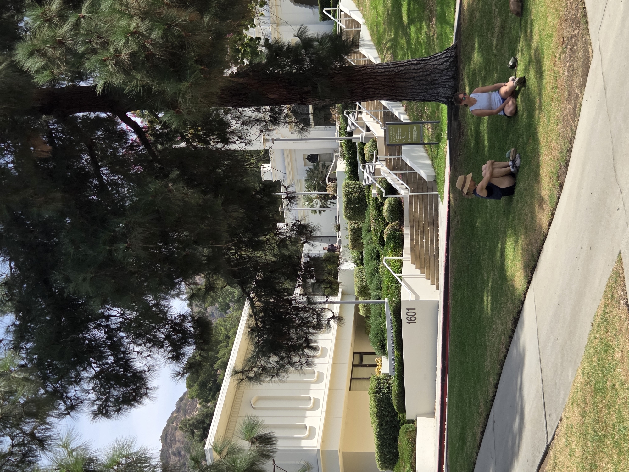 The image shows the Brand Library & Art Center in Glendale, California, on a sunny day. Two women are sitting on the grass in the foreground. The woman on the left is wearing a wide-brimmed hat, a dark tank top, and dark shorts. She is sitting with her legs crossed and her arms resting on her knees. The woman on the right is wearing sunglasses and a white tank top with blue shorts. She is sitting with her legs extended. A leash is visible connecting her to a small brown object on the ground, possibly a dog.
Behind the women, a large pine tree stands prominently, its branches casting shadows. The building of the Brand Library & Art Center is visible in the background, a white structure with cream-colored architectural details and several arched windows. A sign with the text "BRAND LIBRARY & ART CENTER" is posted near the stairs leading up to the building. The number "1601" is visible on a white wall near the stairs. The building is surrounded by manicured hedges and trees, with a hilly, tree-covered landscape in the distance. A paved sidewalk runs along the bottom of the image, bordering a patch of dry grass and the green lawn where the women are sitting.