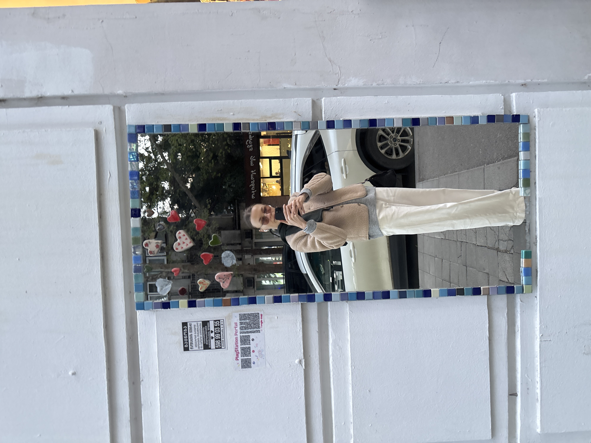 A person is taking a selfie in a mirror with a mosaic tile frame attached to a white paneled wall. The reflection in the mirror shows the person holding a phone, wearing a tan jacket, grey shirt, black mask, and sunglasses. Behind the person, there is a white car with a black tire and a building. To the left of the mirror is a tree decorated with heart-shaped objects. Below the mirror are two posters. The poster on the left contains Georgian text and a phone number, while the poster on the right promotes "PlayStation Portal." Based on the Georgian text and style of the street signs, the location is likely in the city of Tbilisi, Georgia.