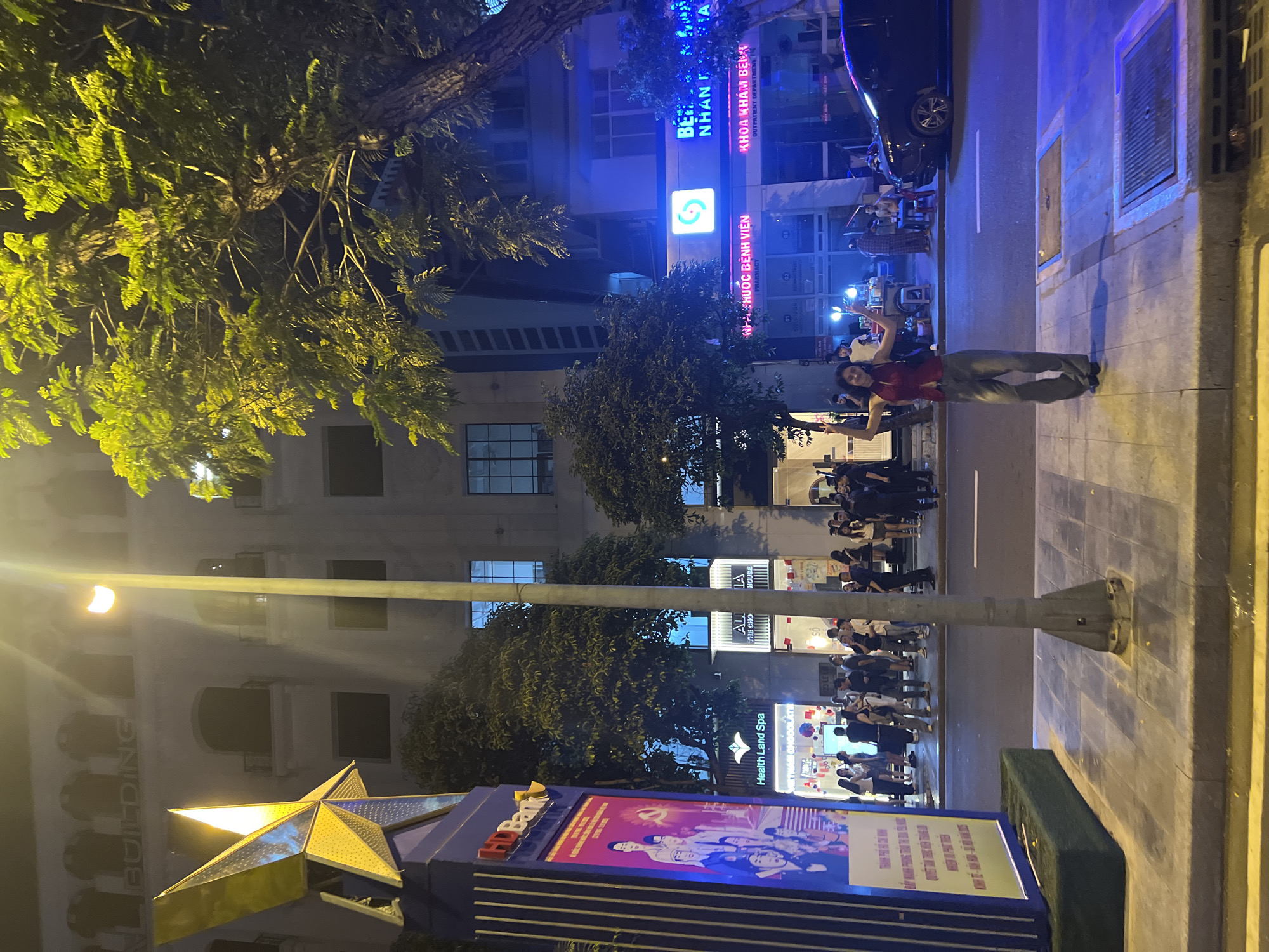 The image shows a nighttime street scene. On the right side of the street, a person is standing. A car is parked nearby. Further down the street, a group of people are gathered. On the left side of the street, there is a brightly lit star sign. The sign also features the words "HDBank". Above the bank sign, there is a building with a sign that reads "Health Land Spa". There are several buildings in the background. The buildings have signs that read "NHÀ THUỐC BỆNH VIỆN PHARMACY", "ALI THE CHO IA HOUSE", and "KHOA KHÁM BENH OUTPATIENT DEPARTMENT". There are trees on both sides of the street.