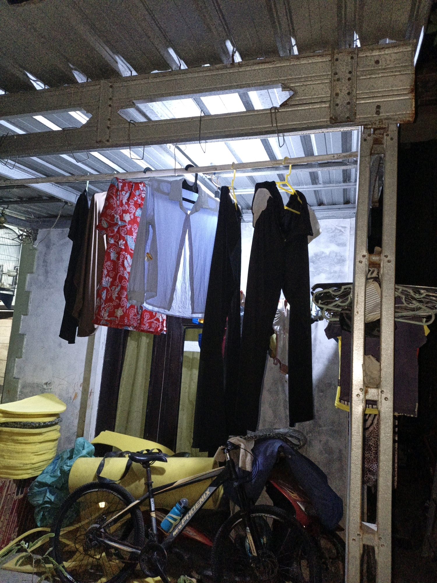 This image captures an outdoor laundry drying area under a corrugated metal roof supported by a galvanized steel frame. Several pieces of clothing are hung on a clothesline, including a red patterned shirt, a light blue long-sleeved shirt, and two pairs of black pants. A bicycle is parked in the foreground, leaning against a stack of yellow foam mats and a blue tarp. The background shows a concrete wall with some visible discoloration and a doorway. The scene appears to be located in a residential area in Cikal, Indonesia, likely a backyard or side alley. The lighting suggests it is either early morning or late evening. There are no people visible in the image.