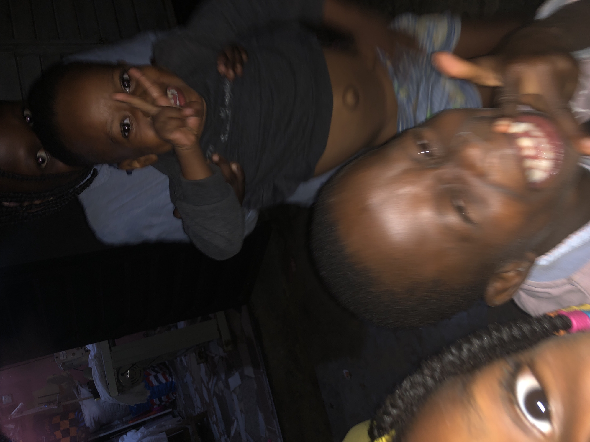 The image depicts a close-up, low-light scene of four young children in an indoor, likely domestic setting. In the foreground, a child's face fills the lower right quadrant, their mouth open in a wide grin, showing teeth. Their right hand is raised, giving a thumbs-up gesture. To the left and slightly behind, another child's eye is visible, looking directly at the camera, with braided hair adorned with colorful beads. Further back, a third child is positioned, wearing a grey long-sleeved shirt and patterned shorts, holding their hands up to the sides. A fourth child is partially visible behind the third, their face obscured. The background appears to be a room with some stacked materials, possibly fabric or goods, and what looks like a sewing machine on a table to the far left. The floor is uneven and appears to be tiled or concrete. The scene suggests a casual, informal gathering, with the children posing for a photo. The overall lighting is dim, with some ambient light contributing to the scene. No explicit indicators of the city of Ado, Nigeria are present in the image itself.