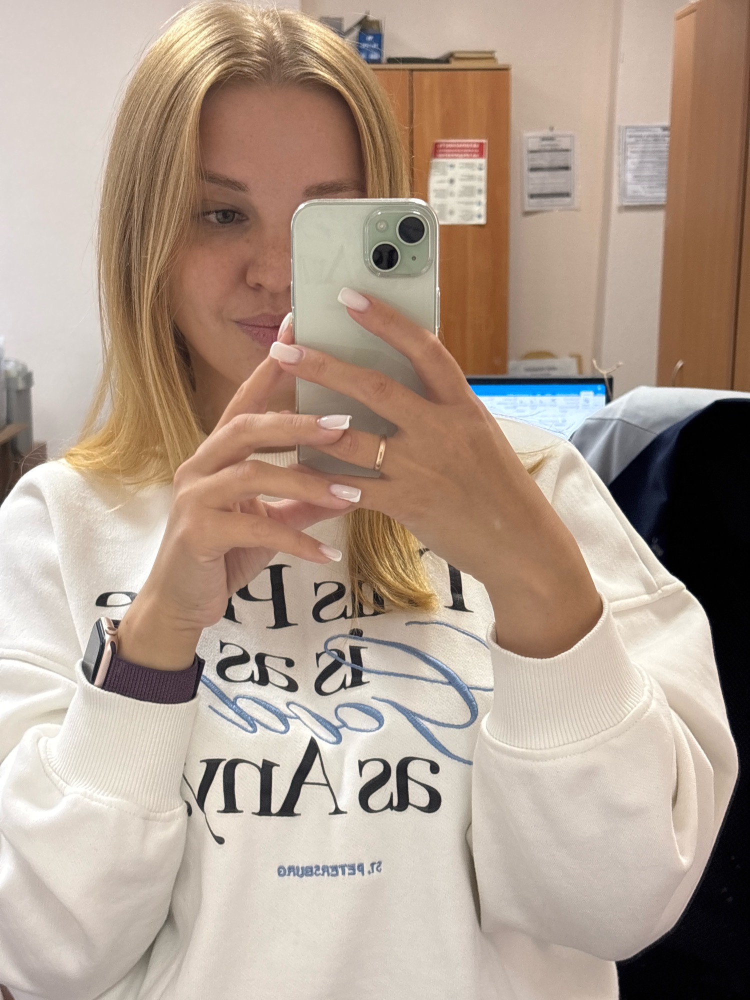 A young woman with blonde hair is taking a selfie in an office environment. She is wearing a white sweatshirt with black and blue text. She is holding a light-colored smartphone in her hands, and her left wrist has a smartwatch with a dark purple band. In the background, there are office cabinets and posters on the wall. The image does not provide enough information to determine the specific location in the city of Rusanovo, Russia.
