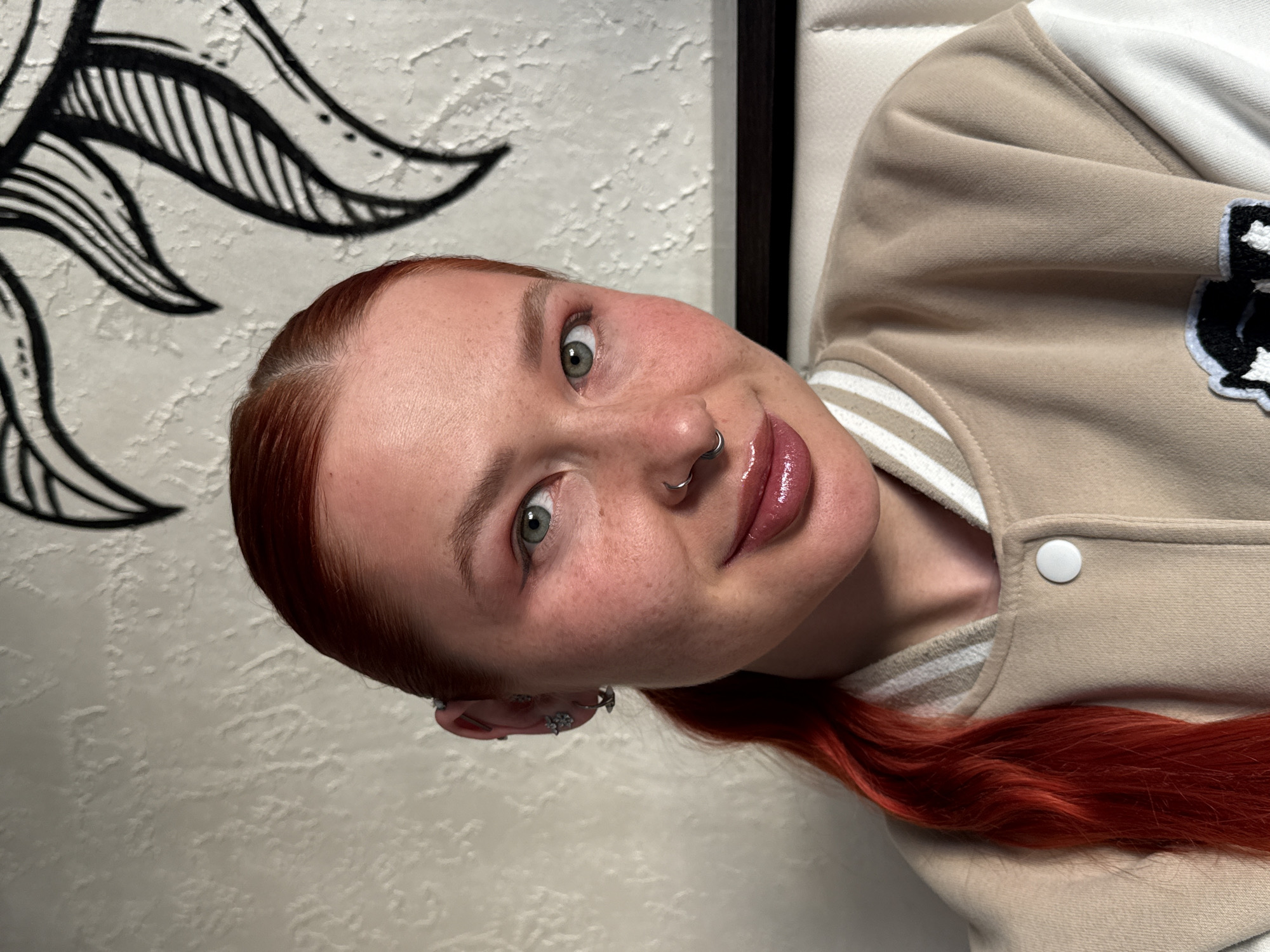 A portrait-style image, rotated 90 degrees clockwise, features a young woman with fair skin, numerous freckles, and striking reddish-orange hair. Her head is tilted, and she looks directly into the camera with wide, light green or hazel eyes. A slight, confident smile plays on her glossy lips. She has a silver septum piercing in her nose and several silver earrings, including a helix piercing and others, visible in her left ear.
She is wearing a casual, light beige jacket or sweatshirt with white accents and what appears to be a black and white patch or emblem on the right side of her chest. White and beige stripes are visible on the cuff of her left arm. A strand of her long, red hair rests across her shoulder, over the jacket.
The background consists of a textured, off-white wall, possibly stucco or a similar finish, adorned with a stylized black and white graphic of a sun or floral design on the upper left. A dark, possibly black, frame or border is visible on the far right side of the image. The setting appears to be indoors. There is no visible text in the image. The general atmosphere is casual and engaging, with the woman's direct gaze conveying a sense of self-assurance. The provided context suggests the location is Rostov-on-Don, Russia, though this is not visually evident in the image itself.