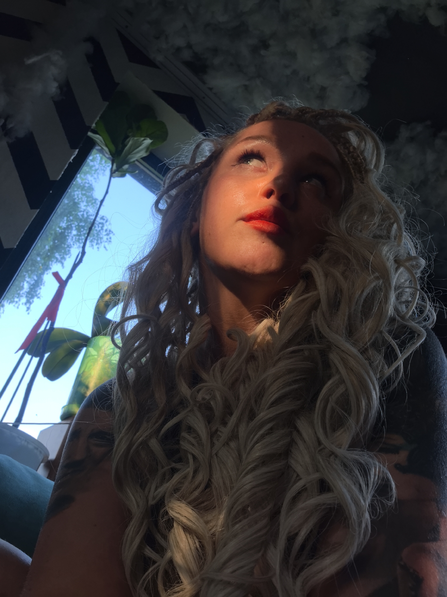 A medium shot captures a woman with blonde, curly hair and braided accents, looking upwards. She is wearing bright red lipstick and has tattoos on her left arm, which is visible. Her face is illuminated by a strong light source from the upper right, casting a dramatic shadow across her cheek. In the background, a window reveals a bright blue sky and trees outside. To the left, a black and white chevron pattern is visible on a wall, along with some white, cloud-like decorations. A large green plant with broad leaves is also present near the window. The scene appears to be indoors, possibly in a room with a window overlooking an outdoor environment. The location is described as Noviy, Russia.