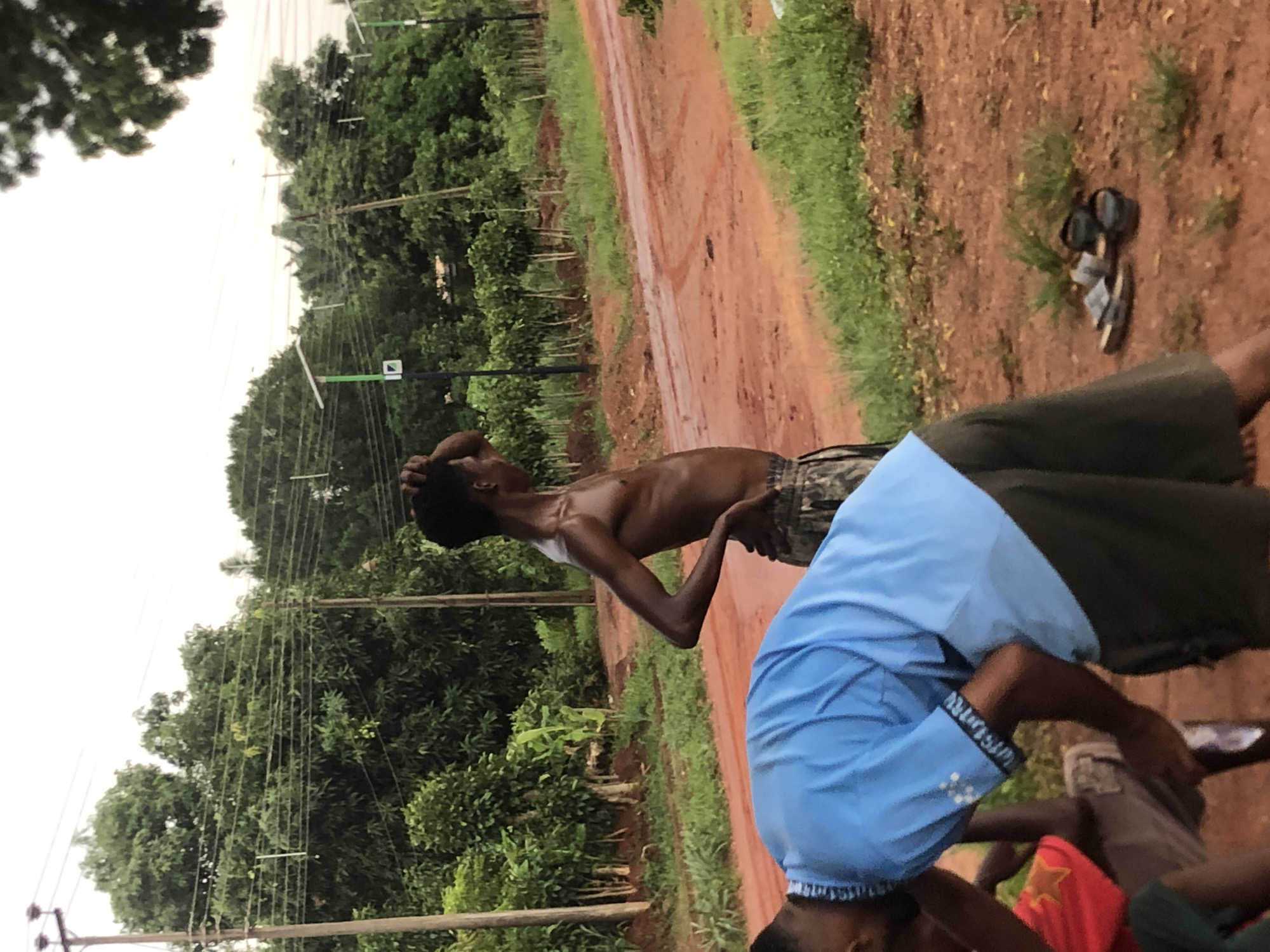 The image, taken in Ukwu-Abwa, Nigeria, captures a quiet outdoor scene in a rural or semi-rural setting. The primary subjects are several individuals along a reddish-brown dirt road that appears damp, possibly from recent rain. Lush green trees and vegetation flank both sides of the road, extending into the background.
In the foreground, part of a person is visible, wearing a light blue short-sleeved shirt with white trim. Text, possibly reading "Wisdom," is faintly visible on the shirt's sleeve. This person is bent over, looking downwards. Further back, a shirtless man wearing camouflage shorts stands upright, looking upwards or into the distance with one hand on the back of his head, suggesting observation or contemplation. A glimpse of a third person in a red shirt with a yellow star can be seen in the bottom right corner.
A pair of black and grey sandals rests on the dirt ground near the shirtless man. Utility poles with numerous power lines run through the scene, vertically dissecting the dense foliage. The sky is overcast, indicating a cloudy day. The overall impression is one of calm and daily life in a verdant, earthy environment.