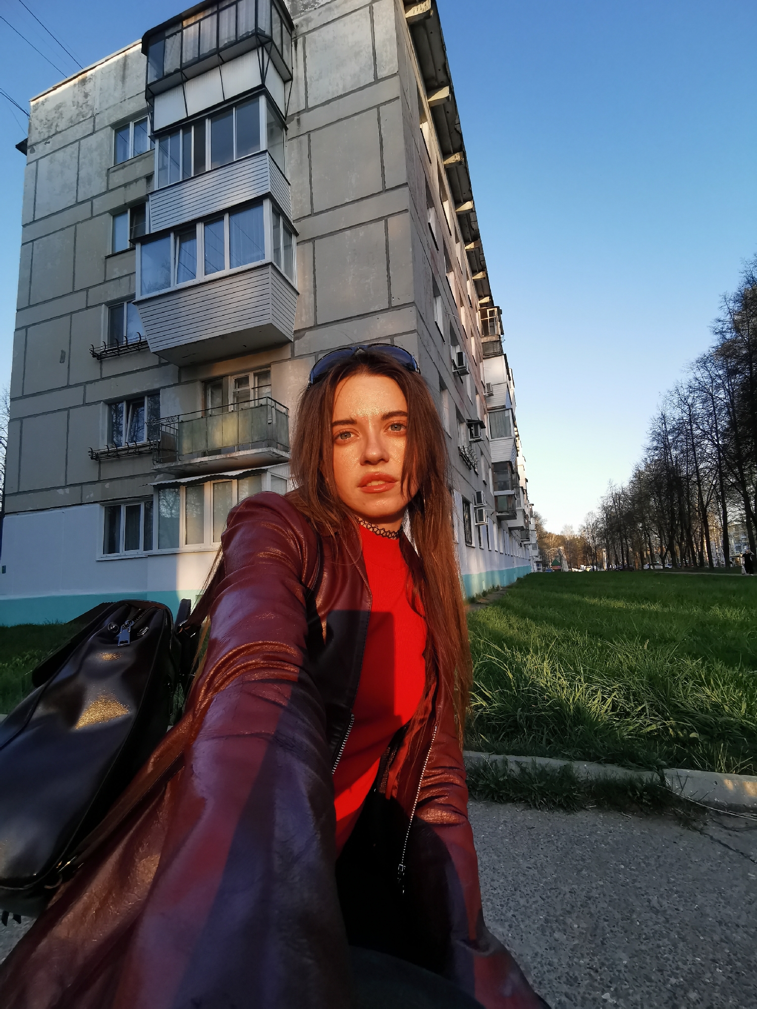 A young woman in a red top and burgundy leather jacket is taking a selfie outdoors in front of a large, gray apartment building. She has long, reddish-brown hair and is wearing sunglasses on her head. A dark leather bag is visible on the left side of the frame. The background includes a grassy area and trees lining a sidewalk. The sky is blue. The photo appears to be taken during the golden hour, with warm sunlight illuminating parts of the scene.
