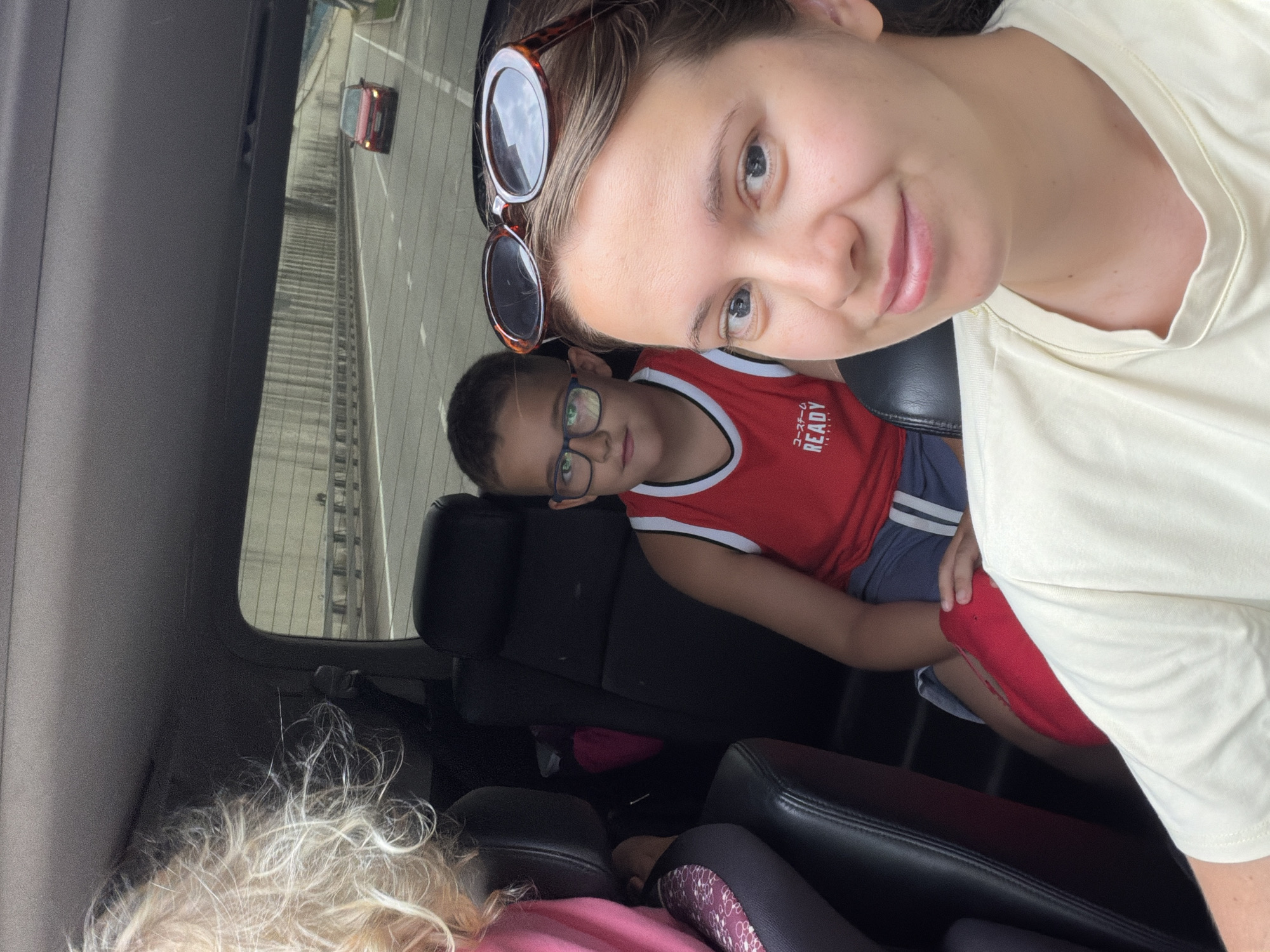 In a frontal selfie taken from inside a vehicle, three people are visible, with a highway visible through the rear window. The person in the foreground is a young woman with light brown hair, wearing a light yellow t-shirt and sunglasses perched on her head. To her left and slightly behind, a young boy with short brown hair and glasses is wearing a red tank top with white trim and "READY" written on it, and gray shorts. Further to the left and out of focus, a person with blonde curly hair is visible. The interior of the car appears to be black leather. Outside the vehicle, a red car is seen driving on a highway with concrete barriers and trees. The scene is set on a highway in Sochi, Russia.