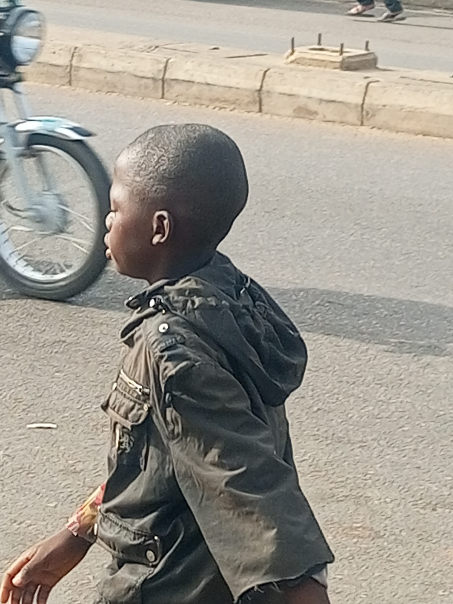 A child is walking on a paved road in what appears to be a city setting. A motorcycle is partially visible to the left. The road has a curb. Another person is walking on the other side of the road. The child wears a dark-colored jacket with a hood. The location is Tempure, Nigeria.
