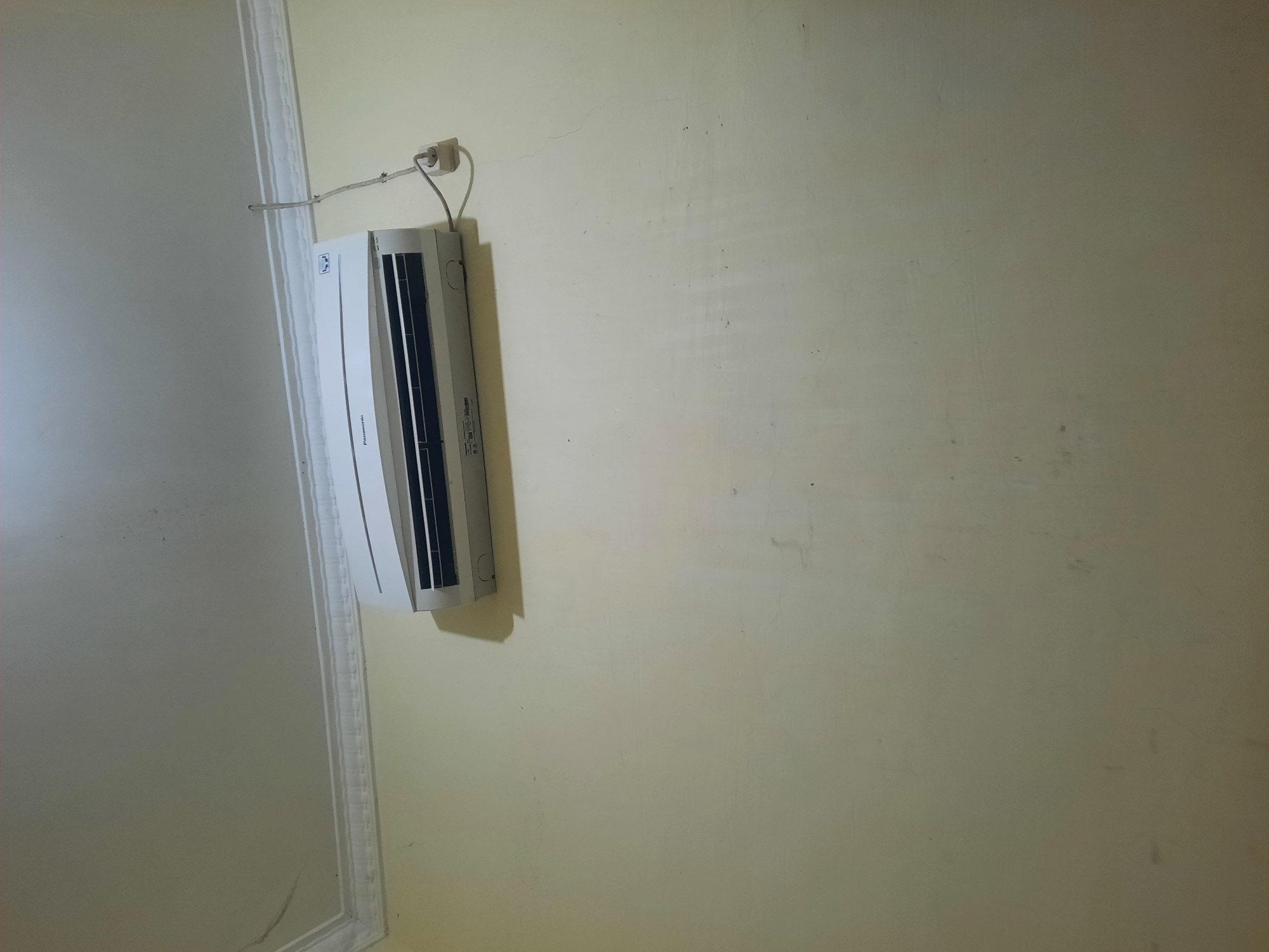 The image shows a split-unit air conditioner mounted on a cream-colored wall beneath a white ceiling with ornate molding. The air conditioner unit is white with horizontal louvers. A grey electrical cord runs from the unit to a white wall socket. The wall appears to have some subtle texture and minor marks. The lighting suggests an indoor environment, likely a room. Based on the provided context, this scene is located in the city of Lhokseumawe, Indonesia.