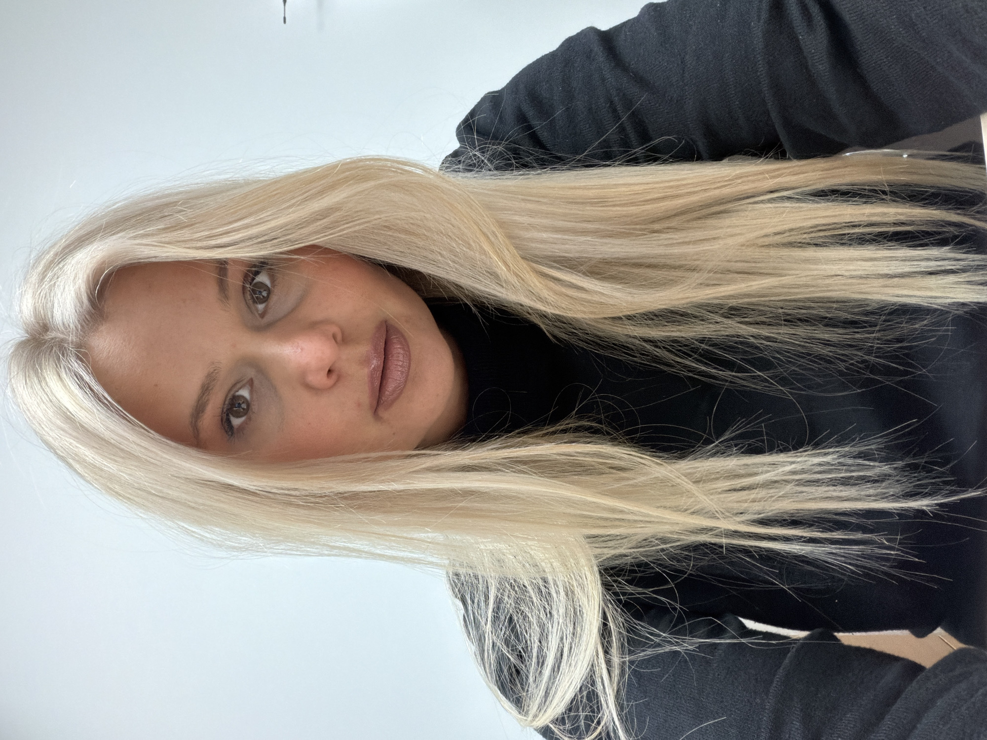 A woman with long, light blonde hair is looking at the camera. She is wearing a black turtleneck shirt. The background is a plain white wall. The image is oriented vertically. There is no indication of location in the city of Sarapul, Russia.