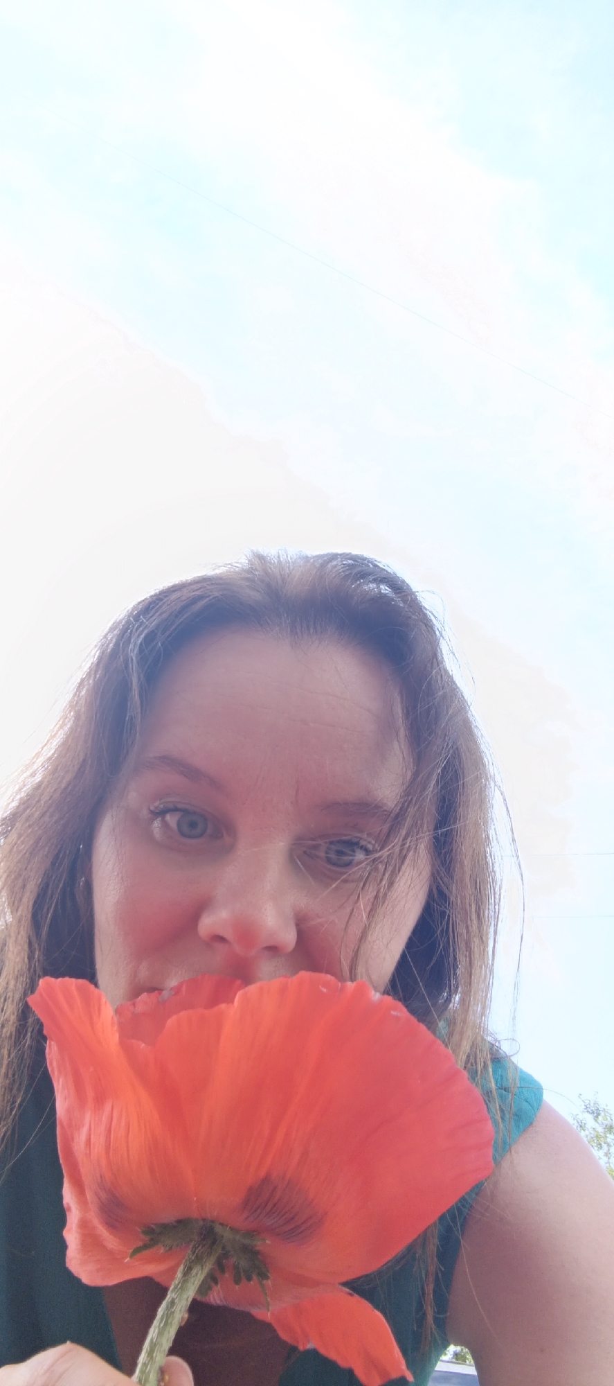 A low-angle, vertical, full-body shot depicts a woman holding a vibrant red poppy flower in front of her face, obscuring her mouth and nose. She has light brown hair and blue eyes, and her head is tilted back as she looks upwards. The background consists of a bright, hazy sky with some light cloud formations. The woman is wearing a dark teal garment. The scene appears to be outdoors on a sunny day, given the brightness of the sky and the lack of artificial lighting. There is no indication of the city of Chaikovsky, Russia, in the image.