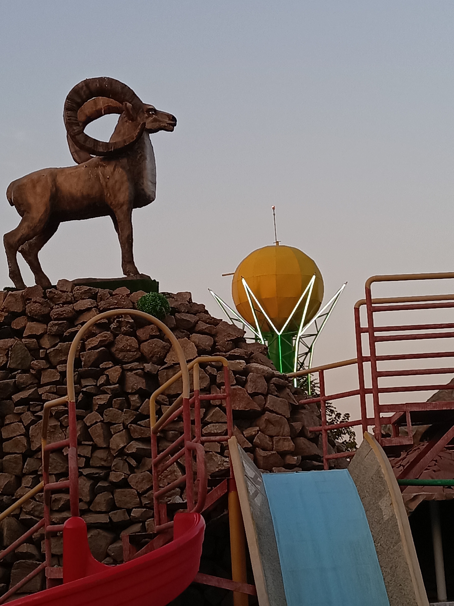 The image displays a statue of a bighorn sheep positioned atop a mound of rocks, with a playground slide in the foreground. In the background, a brightly lit, spherical structure resembling a lotus flower stands against a pale sky. The playground features red and yellow railings and a blue slide. The scene is likely located in Shymkent, Kazakhstan, possibly at an amusement park or recreational area. There are no people visible in the image.