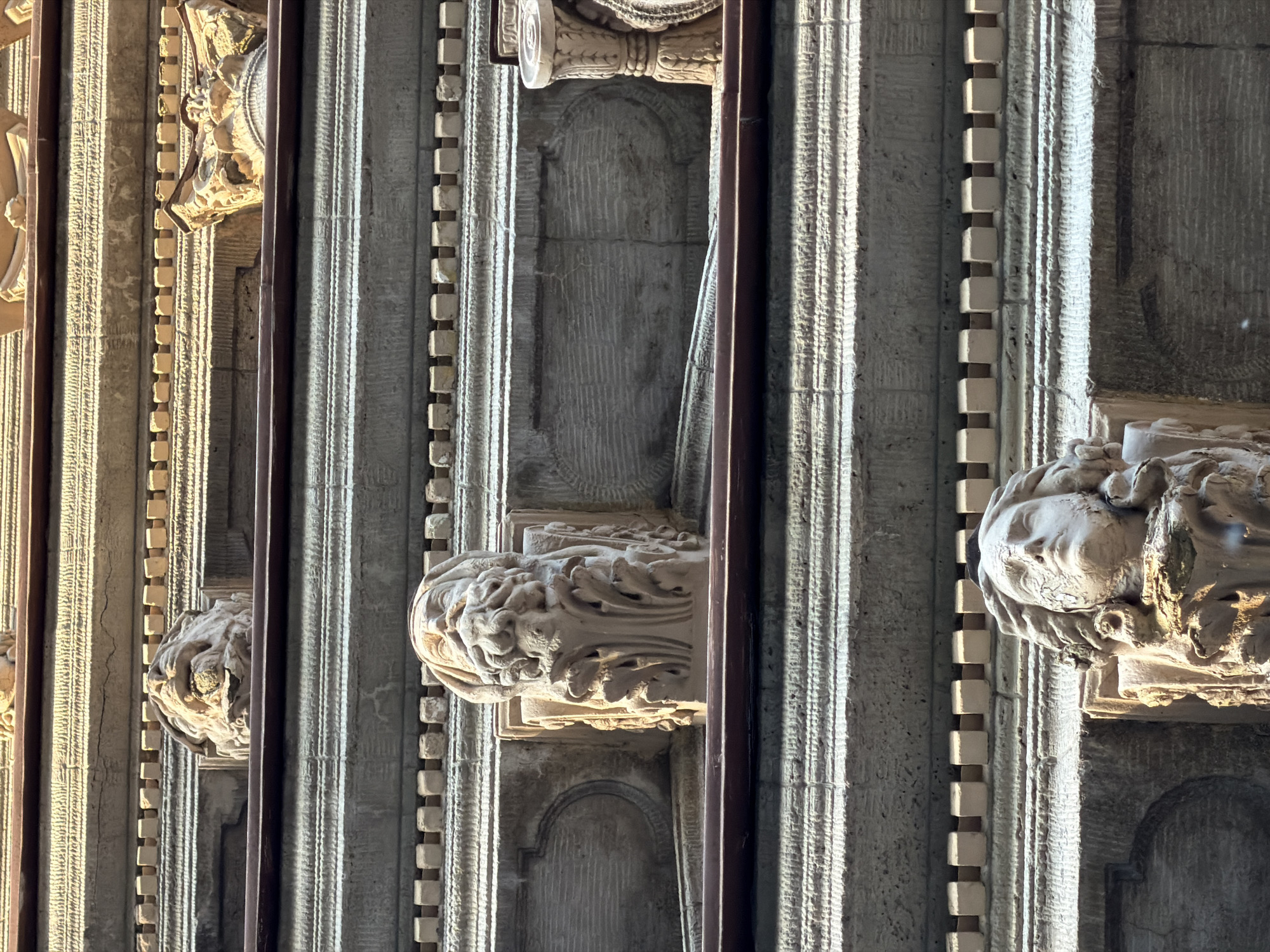The image shows a close-up view of architectural details on the facade of a building. It features several carved stone elements, including grotesque faces and decorative moldings. The architectural style suggests a historical building, possibly from the Baroque or Neoclassical period. The light and shadow patterns indicate it is daytime, with the sun casting harsh highlights and deep shadows on the stone. The facade is made of large, rough-hewn blocks of stone with horizontal banding and decorative friezes. In the foreground, two distinct carved faces are visible. The upper face appears to be a lion or a creature with a mane and open mouth, while the lower face is a human-like figure with a beard and flowing hair, looking upwards. Between these two prominent faces are other decorative elements, including carved columns and recessed panels with textured surfaces. The overall impression is one of weathered, ornate stonework.
This architectural detail is located on the facade of the Singer House (Dom Knigi) in Saint Petersburg, Russia. Specifically, these are decorative grotesques and architectural elements on the building's upper stories.