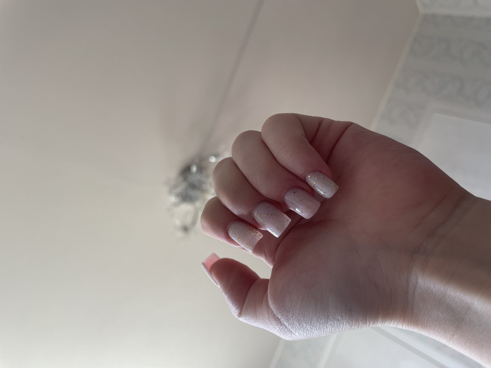 The media file displays a close-up of a person's hand, held up and angled, showcasing a fresh manicure. The hand has light-toned skin, and the nails are of medium length, shaped in a square or soft coffin style. They are painted with a translucent light pink or nude polish, heavily embellished with small, reflective gold glitter flakes that catch the light.
The photograph is taken from a low angle, looking upwards towards an indoor ceiling and wall. The background is largely out of focus, emphasizing the hand and nails. The ceiling, which dominates the upper left portion of the image, is a plain, light color, likely white or off-white. Towards the upper center-right, a blurred, decorative light fixture or chandelier with metallic or crystal elements is visible, hanging from the ceiling. On the far right, a wall featuring a subtle, light-toned patterned wallpaper is faintly discernible, also out of focus.
The overall lighting is soft and ambient, suggesting an indoor setting, possibly illuminated by indirect natural light or artificial lighting. There is no specific activity taking place other than the display of the manicured hand. No text is visible in the image. The scene is set in an interior location in Ольховка, Russia.