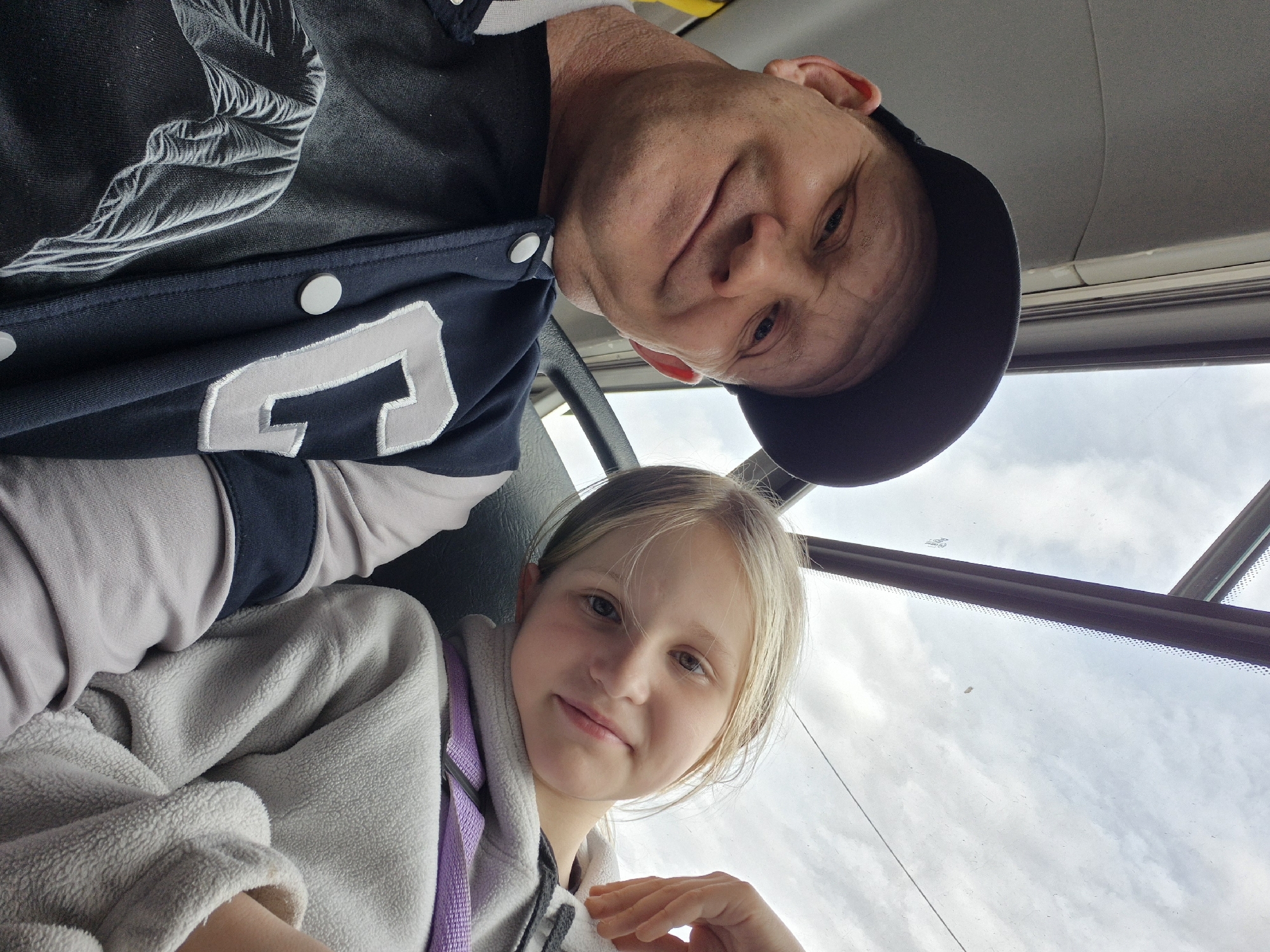 A low-angle photograph shows an adult male wearing a black baseball cap and a dark jacket with a large white "2" on the front, positioned above and to the left of a young girl wearing a light-colored hooded fleece jacket with a purple strap, possibly a backpack strap. The background shows a window with a view of clouds and sky. The photograph appears to be taken from inside a vehicle, possibly a bus or car. The specific location within the city of Chaikovsky, Russia cannot be determined from the image.