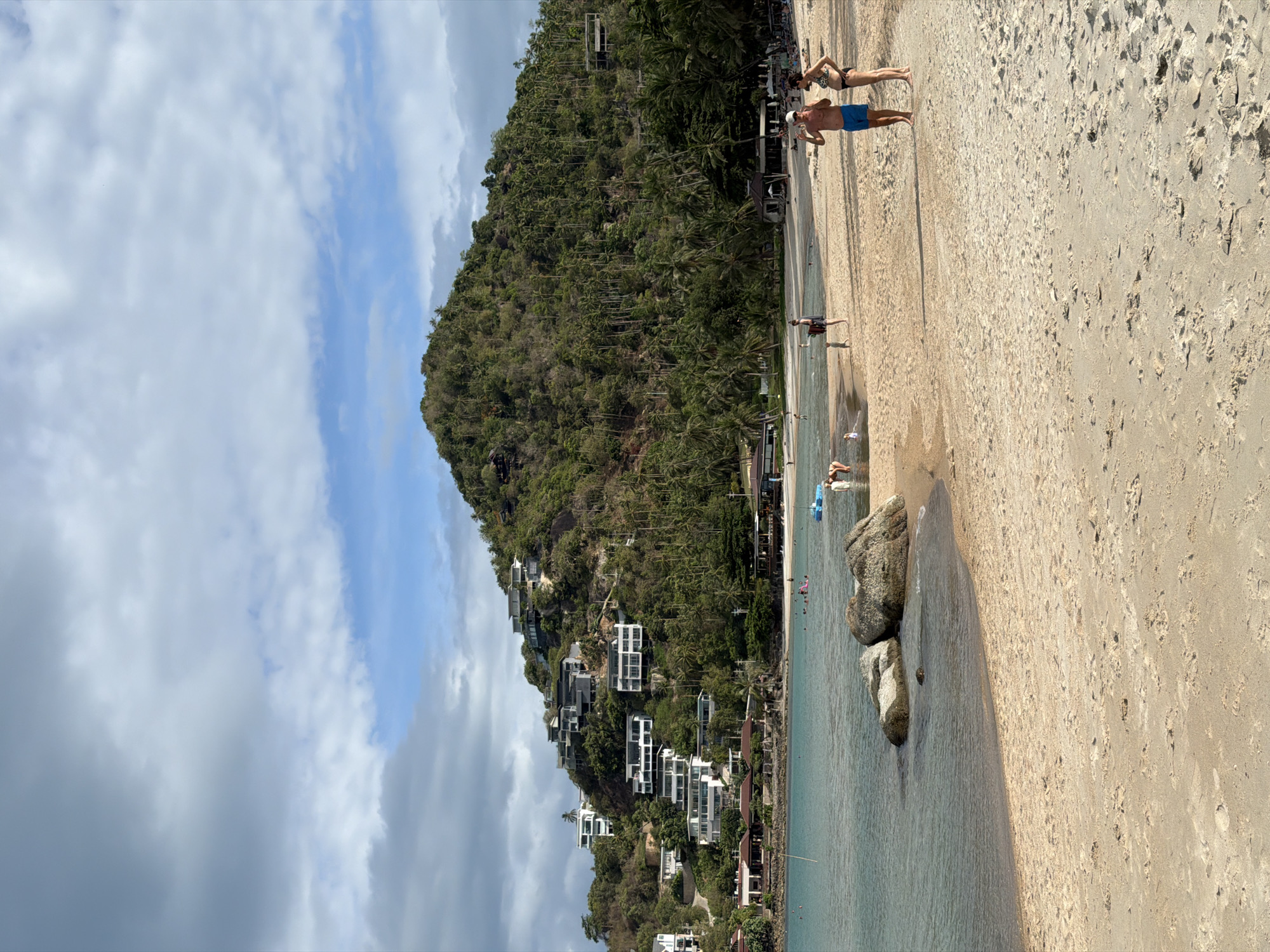 The image shows a beach scene in Baan Chaweng Noi, Thailand. The beach has tan sand and light blue water. Two large rocks are at the edge of the water. People are standing and walking on the beach and in the water. A hill covered with lush green vegetation rises sharply from the beach. White buildings are built into the hillside, some looking like multi-story homes or resorts. The sky is partly cloudy with patches of blue.