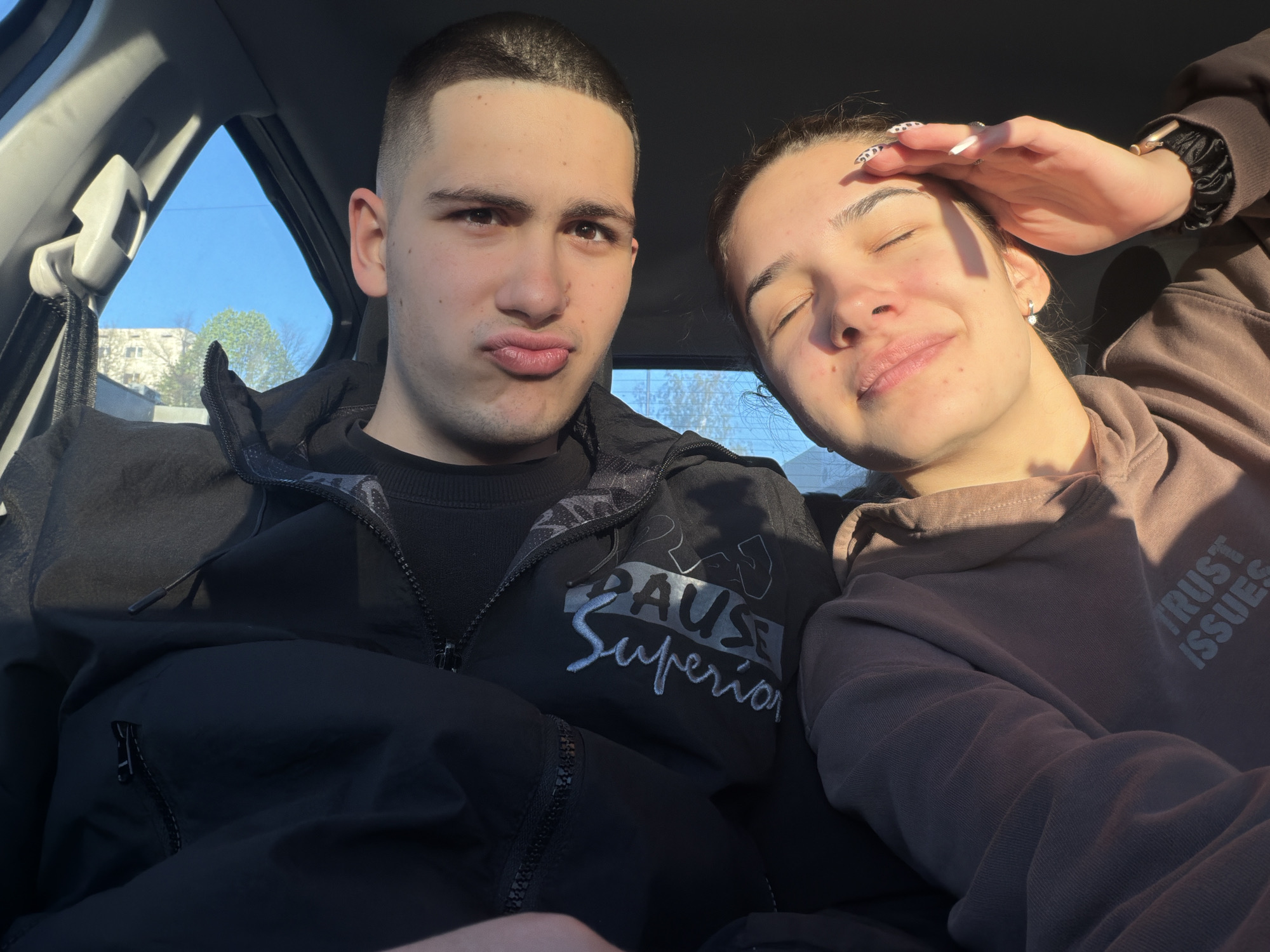 Two young adults are seated inside a car. The male, on the left, has short dark hair, light skin, and is making a pouting expression. He is wearing a black jacket with white text that reads "PAUSE Superior". The female, on the right, has dark hair tied back and is partially shielding her eyes with her hand. She is wearing a brown hoodie with the text "TRUST ISSUES" visible. The background shows the interior of the car, including the window, and glimpse of a building and a tree outside the car window. The location is Sofia, Bulgaria, as indicated by the building exterior and surrounding trees.