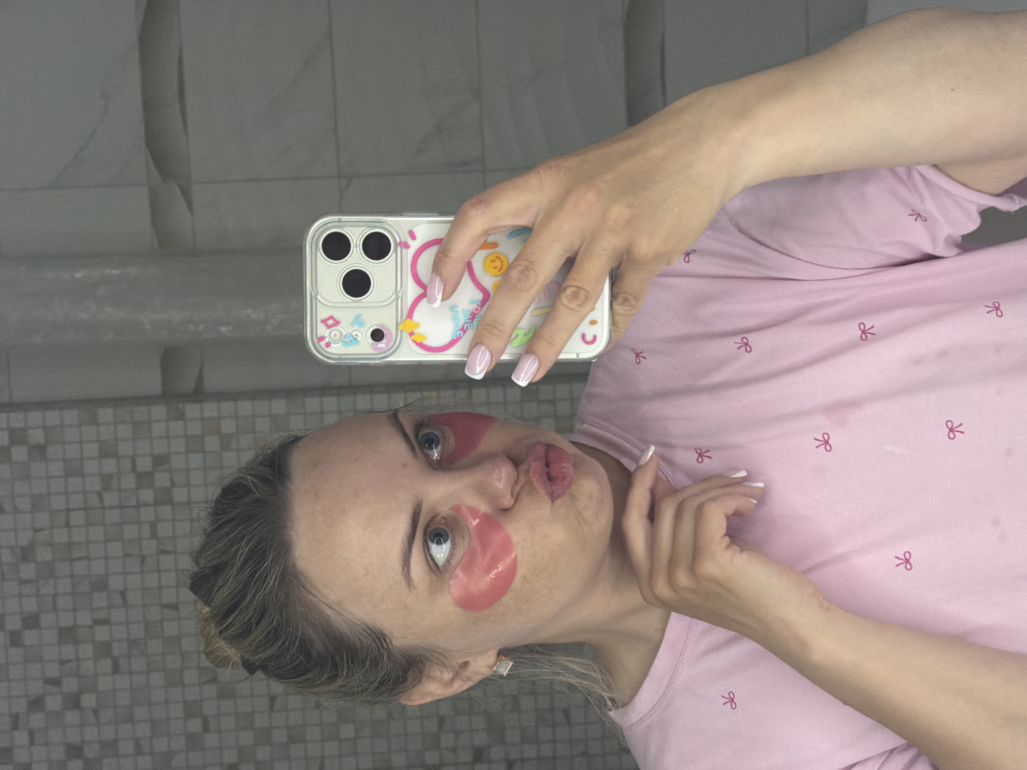 A woman is taking a selfie in a bathroom, likely in an apartment or house, given the tile work on the wall. She has eye patches on, is wearing a pink shirt with bow designs, and has her hair pulled back. She's holding a cell phone with a decorative case, displaying it towards the camera. The background appears to be a tiled wall. There is no immediate evidence suggesting the location is Votkinsk, Russia; this information is not verifiable from the image.