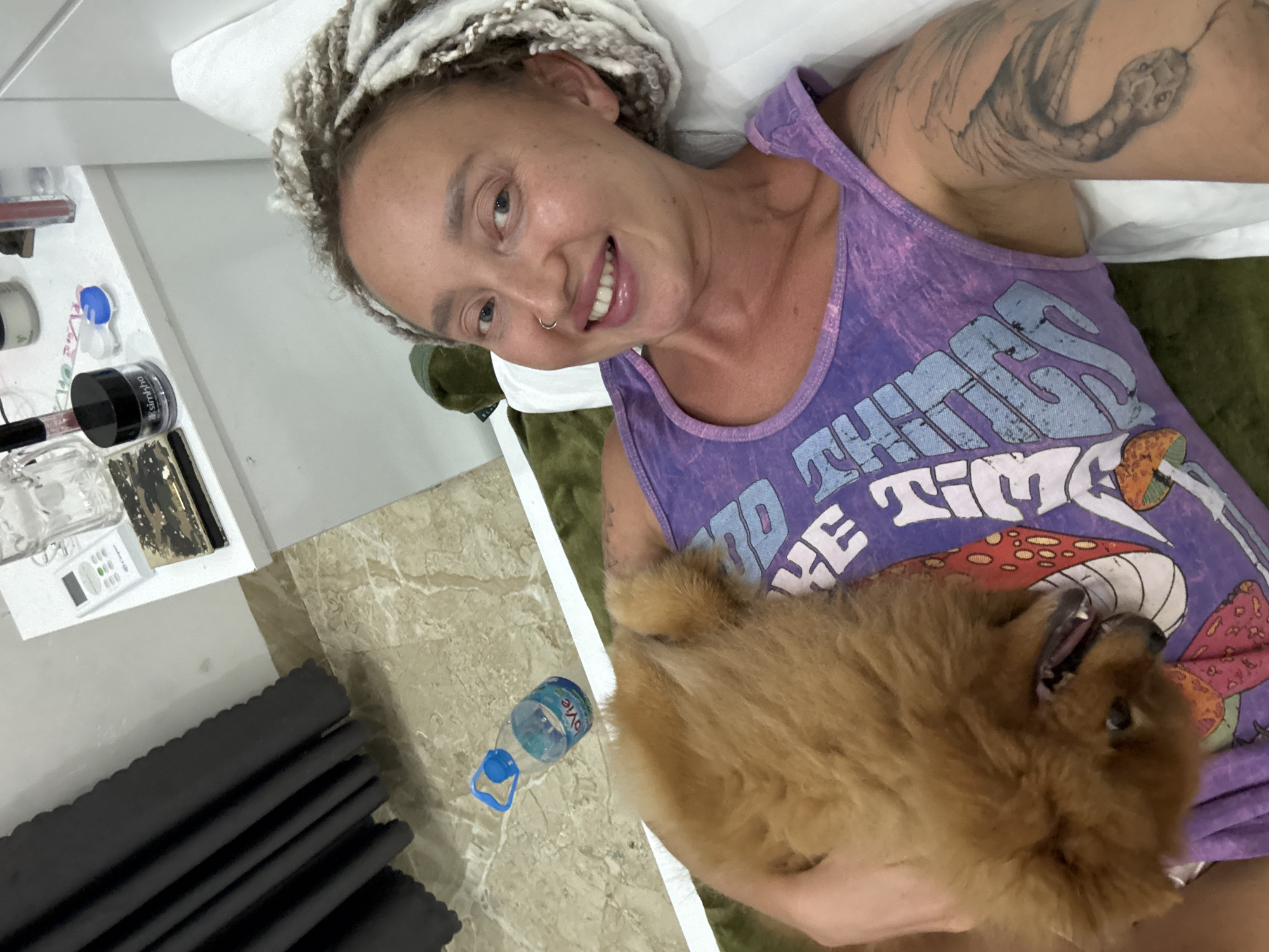 A woman with braided hair and a nose ring lies in bed with a fluffy brown dog. She is wearing a purple tank top with a graphic print. Behind her, a bedside table holds a remote control, a glass, and various cosmetic items. A bottle of Evian water is on the floor next to the bed. The scene appears to be in a bedroom in the city of Bac Nha Trang Ward, Vietnam.