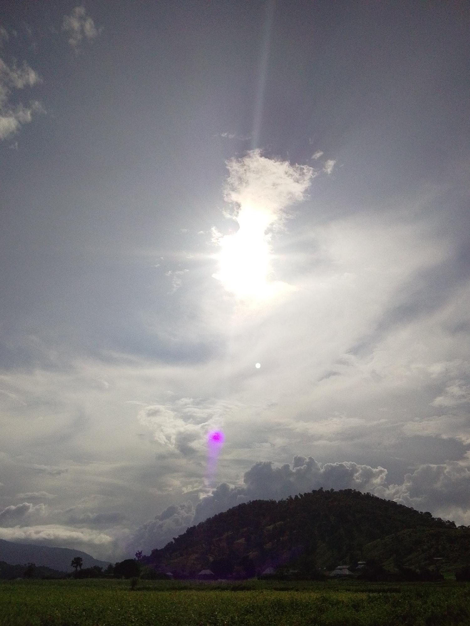 The image depicts a landscape scene in Serti, Nigeria. The sky dominates the upper portion of the frame, filled with a mix of clouds and bright sunlight. The sun is a prominent feature, with rays of light streaming outwards, partially obscured by clouds. There are visible lens flares and light artifacts in the sky, including a bright white spot and a purple vertical streak.
Below the sky, a lush green field stretches across the foreground, suggesting agriculture. In the mid-ground, a dark, tree-covered hillside rises prominently, its silhouette defined against the lighter sky. Scattered houses or structures can be seen nestled at the base of the hill. In the distance, fainter, layered hills recede into the background, adding depth to the landscape.
The overall impression is of an outdoor scene, likely in the late afternoon or early morning given the quality of light. The weather appears to be partly cloudy. There is no visible text in the image. The scene evokes a sense of natural beauty and rural life.