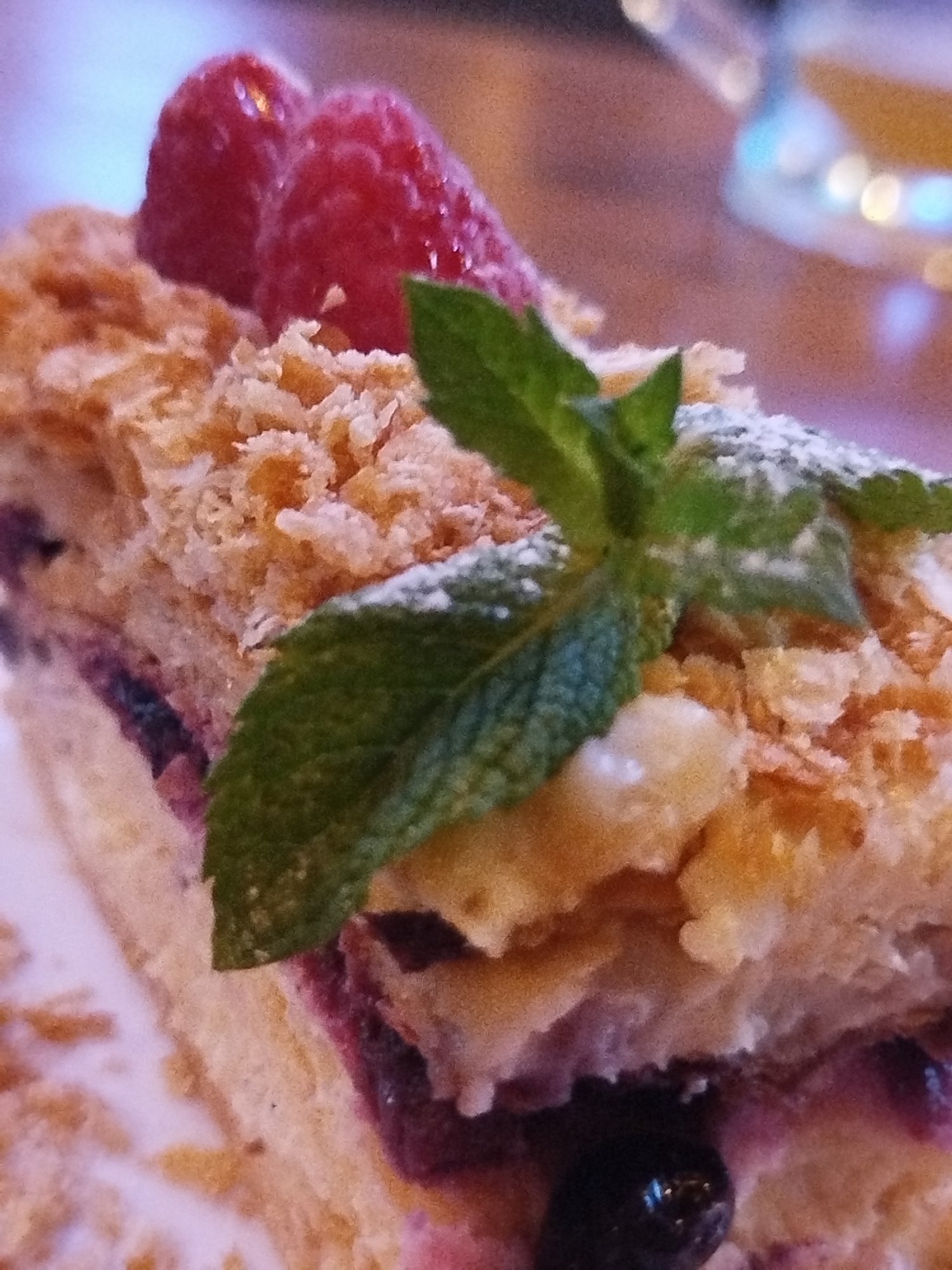 A close-up, eye-level, slightly overhead shot shows a slice of a layered dessert. The dessert has multiple flaky pastry layers, with cream or custard filling between them. The top layer is topped with two raspberries and a sprig of mint dusted with powdered sugar. There are visible blueberries between some of the pastry layers. The background is blurred and appears to be an indoor setting with soft lighting. There is no identifiable signage or landmarks to pinpoint a location in Kremenchuk, Ukraine.