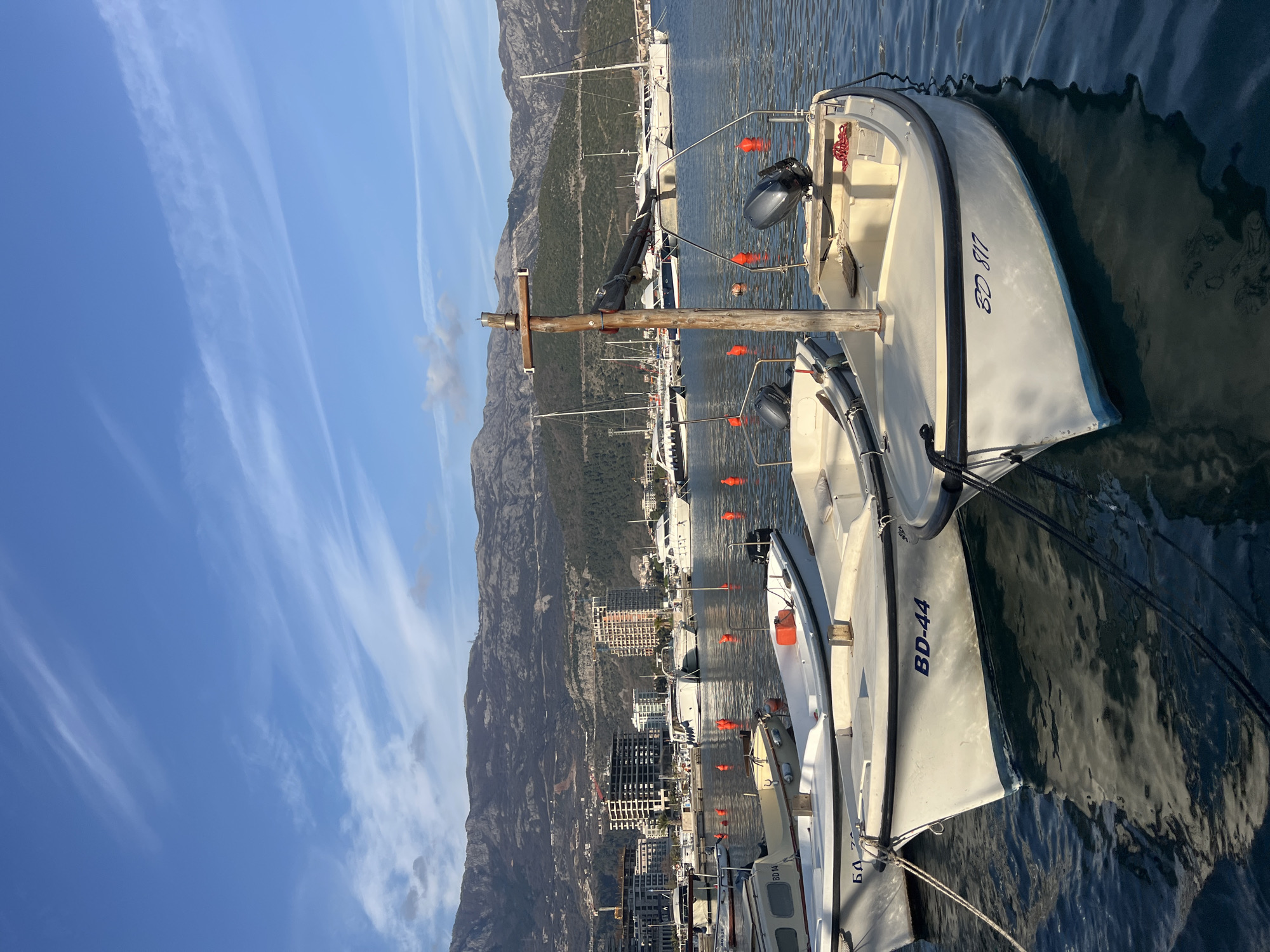 The image depicts a marina scene in Budva, Montenegro, under a clear blue sky with wispy white clouds. The foreground features three small, light-colored boats moored in calm water. The closest boat, on the right, displays "БД 817" on its bow, has an outboard motor mounted on its stern, and features a vertical wooden mast with a horizontal crossbeam. The boat immediately to its left is marked "BD-44" on its bow and also has an outboard motor. A third boat, partially visible on the far left, has "БД 14" on its bow. All three boats have a blue waterline stripe. Mooring lines are visible connecting the boats.
In the midground, numerous larger white yachts and boats are moored in the marina. Several orange spherical buoys are scattered across the water. The background reveals the cityscape of Budva, characterized by multiple multi-story buildings, some appearing modern or under construction, situated along the waterfront. Behind the city, a range of rugged, grey-green mountains rises prominently, forming the distant horizon. Sunlight illuminates the scene, suggesting either morning or late afternoon. No individuals are present in the image.