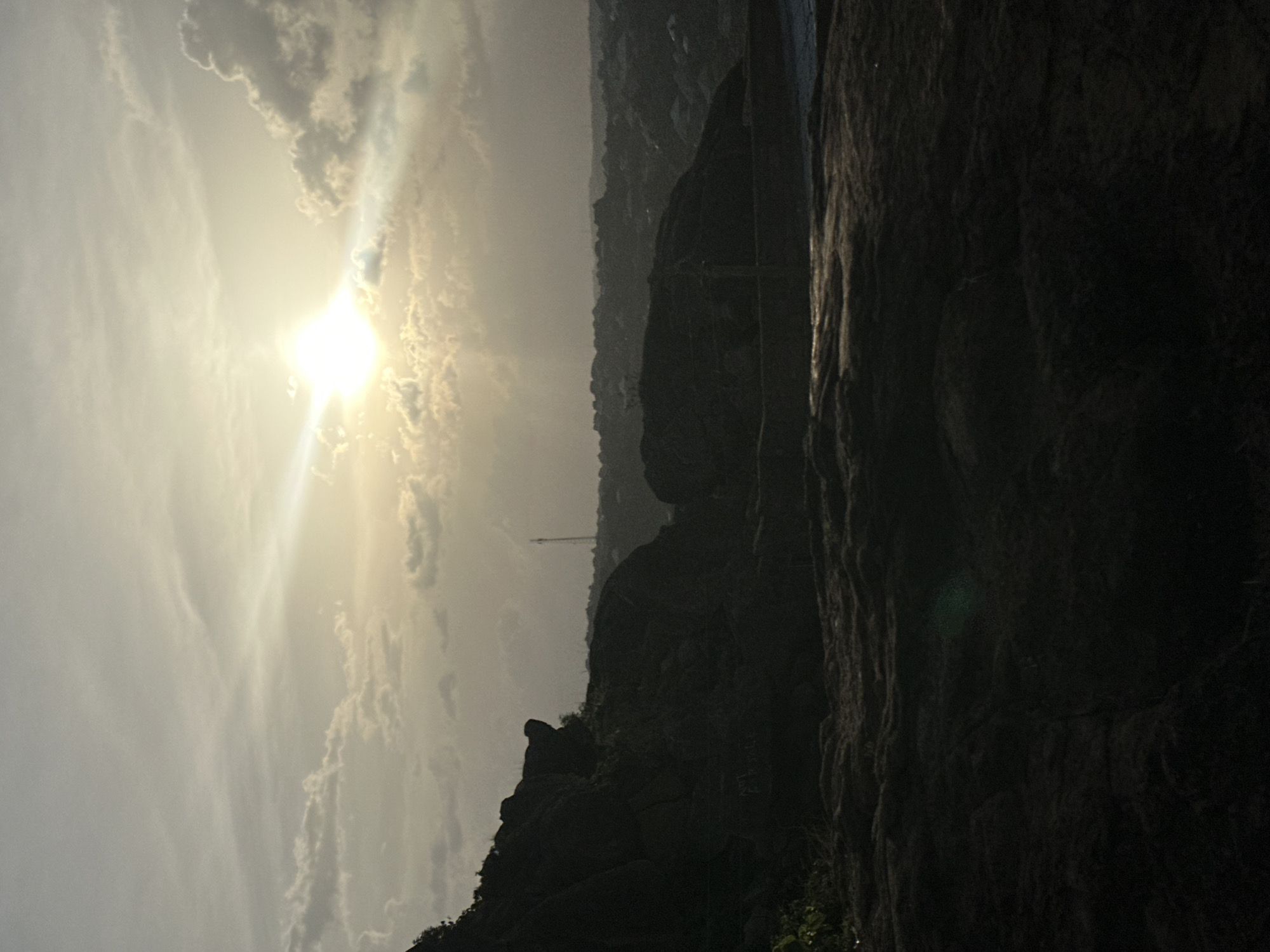 The image displays a dramatic rocky landscape, likely captured during sunrise or sunset due to the intense backlighting. The scene is dominated by a powerful sun on the left, which is a brilliant, almost blinding light source, partially veiled by clouds but casting strong, radiant beams across the sky. The sky itself is filled with a mix of bright, broken clouds and softer, hazy formations, creating a striking contrast with the darkened terrain below.
In the foreground and midground, imposing, dark rock or mountain formations rise steeply, their rugged textures and contours rendered in stark silhouette. These massive landforms occupy a significant portion of the lower and right side of the frame. In the distant background, a series of fainter, hazy hills or mountains are visible, also appearing as dark silhouettes against the lighter horizon. A slender, tall structure, possibly a mast or communications tower, can be faintly discerned atop one of these distant hills.
No people or specific activities are visible in the scene. The weather appears partly cloudy with clear sun. The location cues, especially the prominent rock formations, align with the hilly and rocky terrain often found in the Jos Plateau region of Nigeria. The overall impression is one of natural grandeur and dramatic lighting. No visible text is present.