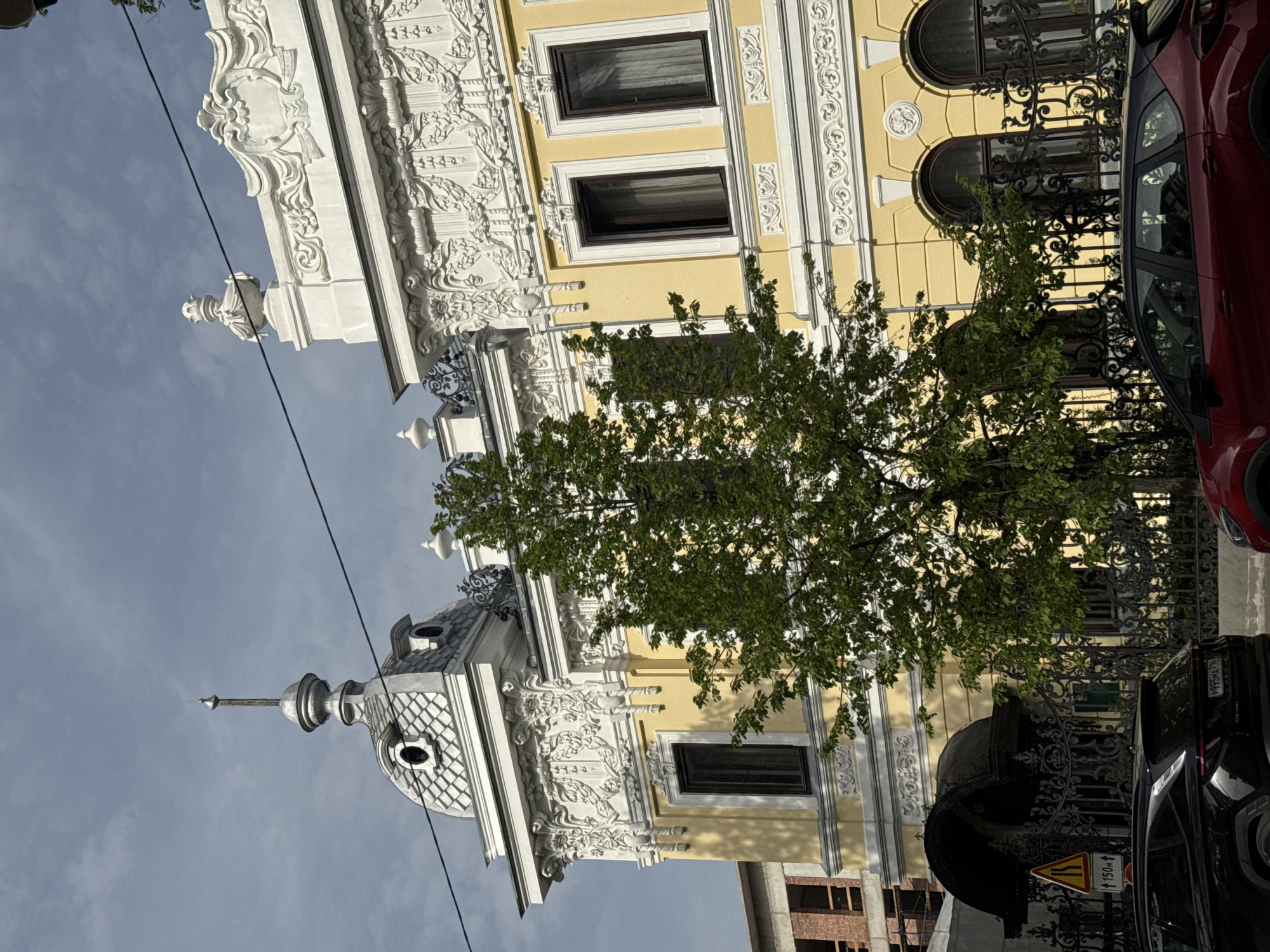 The image depicts a building in Kazan, Russia. The facade of the building is yellow with white decorative elements, including intricate moldings and carvings. There are rectangular windows with dark frames and arched windows with decorative iron railings. A tree with green leaves partially obscures the building. A red car is parked on the right side of the frame, and a black car is parked in front of the building. A yellow and black road sign is visible near the black car. Overhead power lines are visible against the blue sky. The building has a distinctive turret with a pointed spire and a checkered dome.