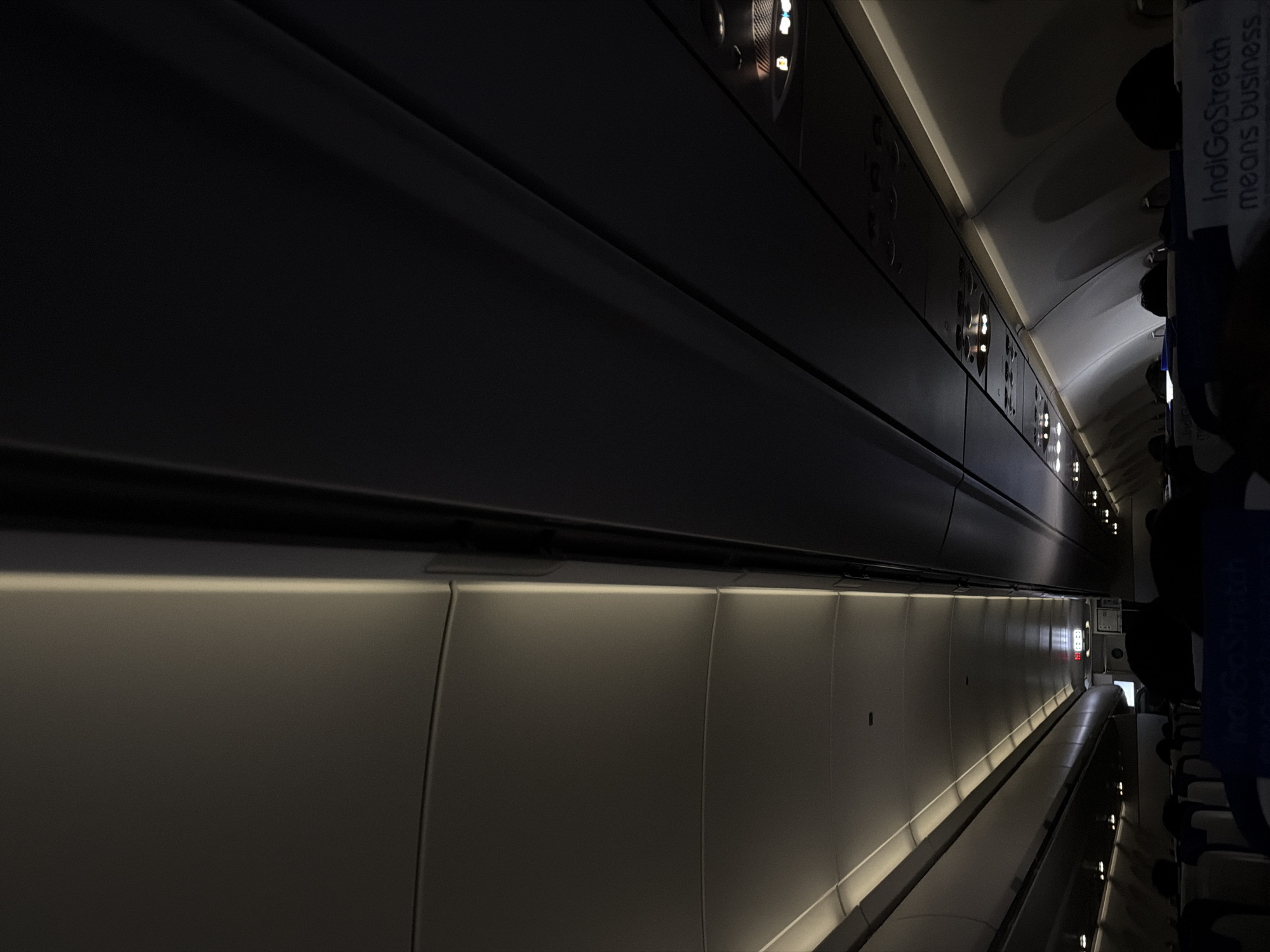 The image depicts the interior of an airplane cabin, likely during a flight as suggested by the dim lighting and overhead compartments. The view is angled upwards, showing the ceiling panels and the rows of seats below, partially obscured by darkness and the heads of passengers. The cabin appears to be illuminated by soft, indirect lighting along the edges of the ceiling panels and some individual reading lights are visible. The colors are muted, dominated by shades of gray and black, with some hints of blue from the seat upholstery. The overall impression is one of a quiet, possibly night-time flight. The text "IndiGoStretch means business" is visible on the headrest covers of the seats in the foreground, indicating the airline. The location in the city of Mamidipally, India, is not directly discernible from the visual elements of the image itself, but it is inferred as the context of the photograph.