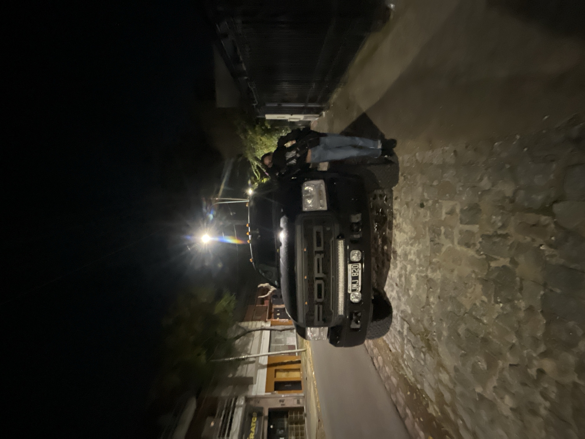 At night, a black Ford truck is parked on a street with buildings on the left side and a wall on the right. A person is sitting on the hood of the truck. The license plate of the truck reads "NMJ 820." The scene appears to be in a residential area. Based on the street signs and architectural style, the location is likely in the city of Olivos, Argentina.