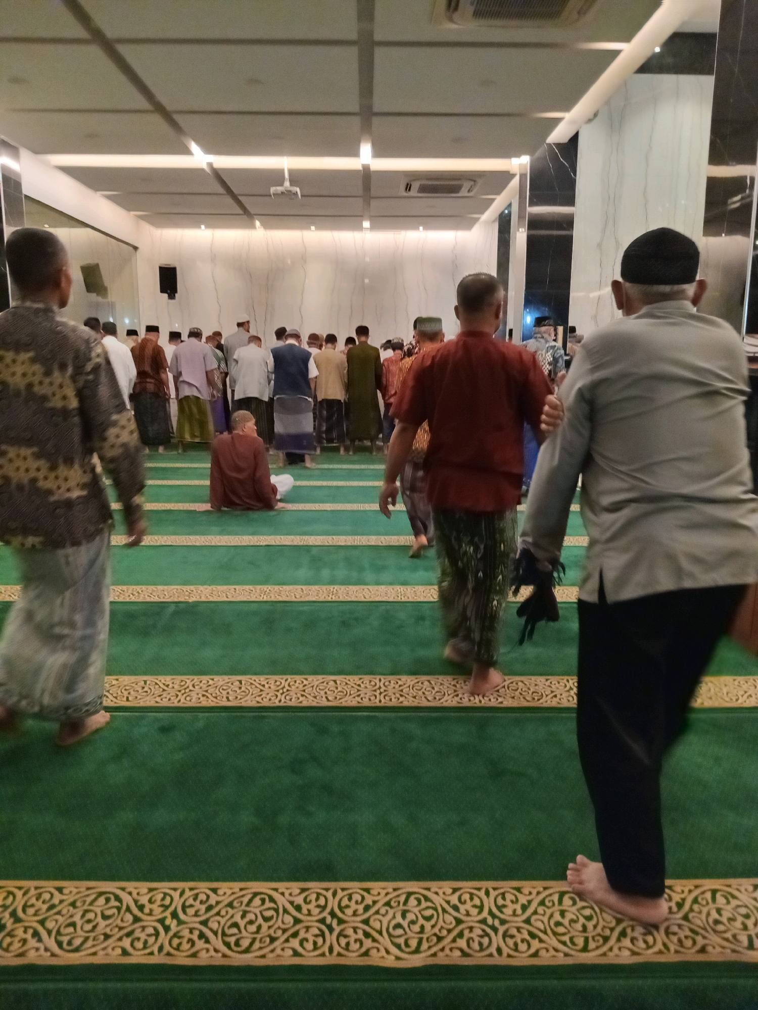 The image shows a group of men in a prayer hall, likely at a mosque in Randuagung, Indonesia. The men are dressed in traditional Islamic attire, including sarongs and shirts. Most of them are facing the front of the hall, suggesting they are participating in a prayer service. The prayer hall has green carpeted floors with decorative gold borders, and the ceiling is illuminated with modern lighting fixtures. In the foreground, one man is seen from behind, wearing a grey shirt and black pants, walking towards the right of the frame. Another man in a patterned shirt and light-colored sarong is walking away from the camera. The overall atmosphere appears to be solemn and respectful.