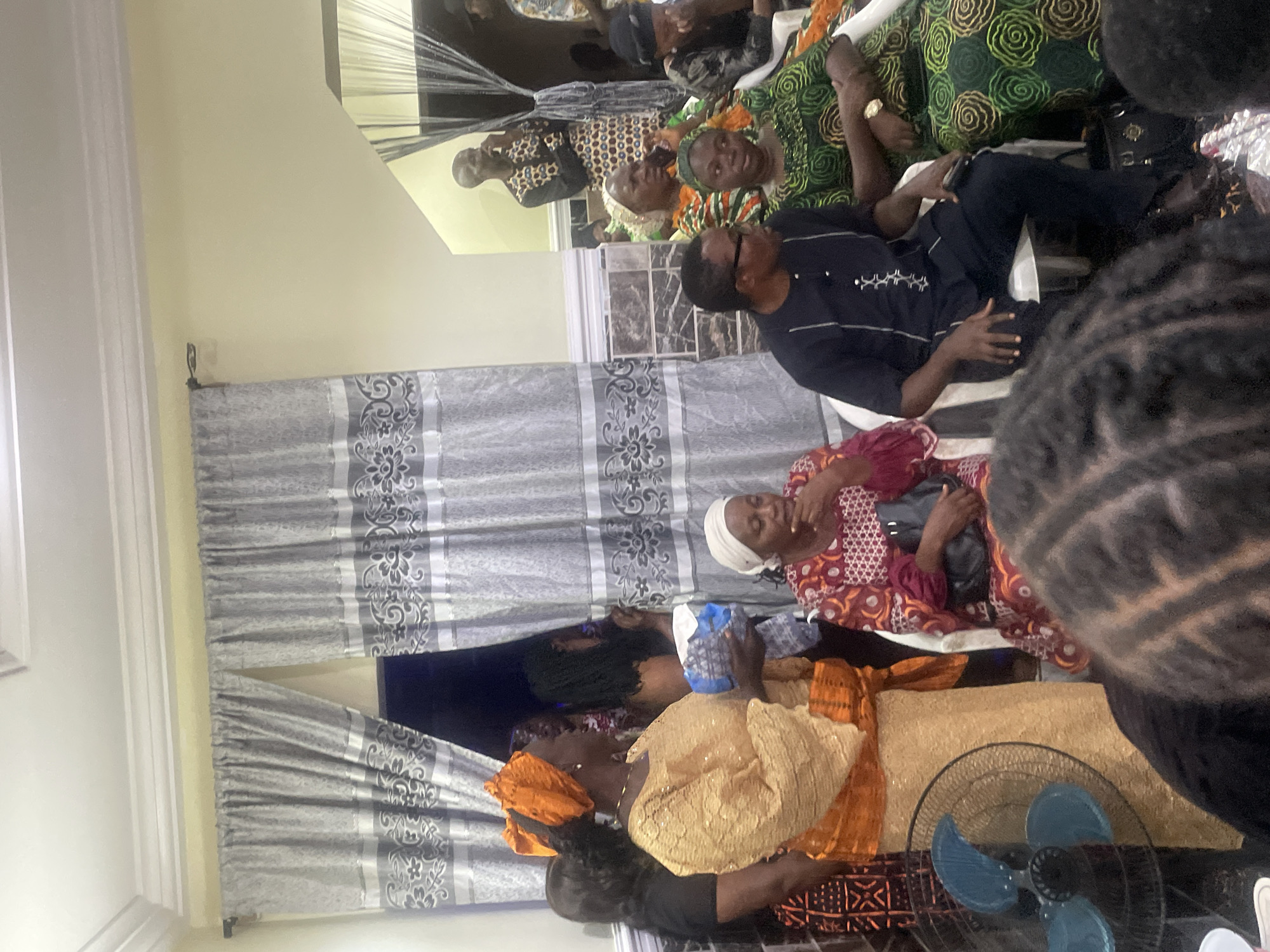 The image depicts a gathering of people, likely at an indoor event in Rukuba Road, Nigeria. The attendees, dressed in traditional African attire, are seated and standing, engaged in conversation and observing something outside the frame. The room is decorated with grey patterned curtains, and a fan is visible in the foreground, suggesting a warm climate. The overall atmosphere appears to be one of community and celebration.
