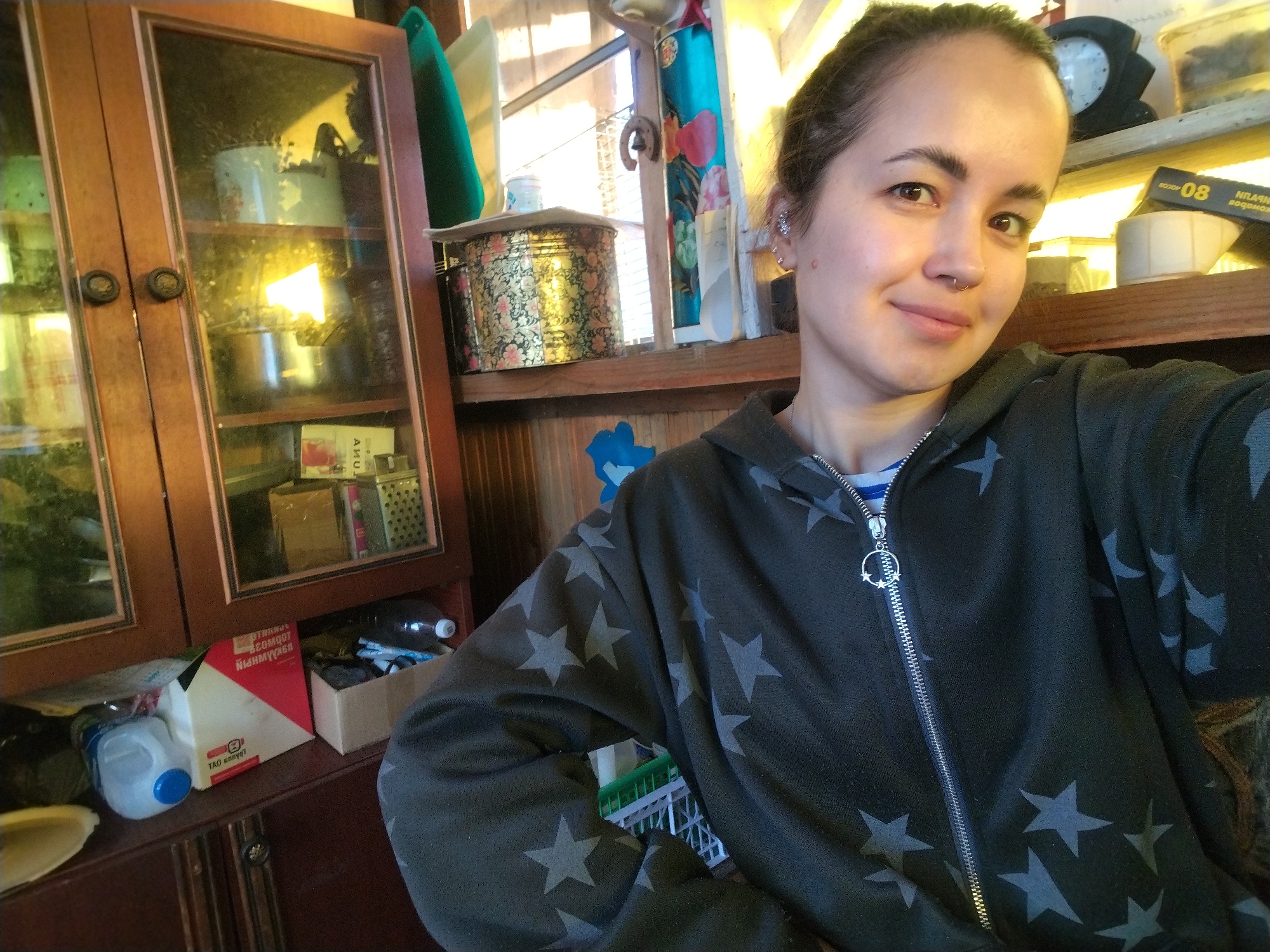 A selfie shows a woman with her hair pulled back, wearing a dark gray hoodie with light gray stars, and with multiple piercings in her ear and a septum piercing. She is smiling slightly at the camera. Behind her is a cluttered interior, including a wooden cabinet with glass doors filled with various items, and shelves with containers, boxes, and a clock. The backdrop also features wood paneling and a window. A partial view of a white and green container is visible on the bottom left and an open box with a bottle inside. The setting appears to be indoors, likely a home. There is nothing in the picture that points to its location being Noviy, Russia.
