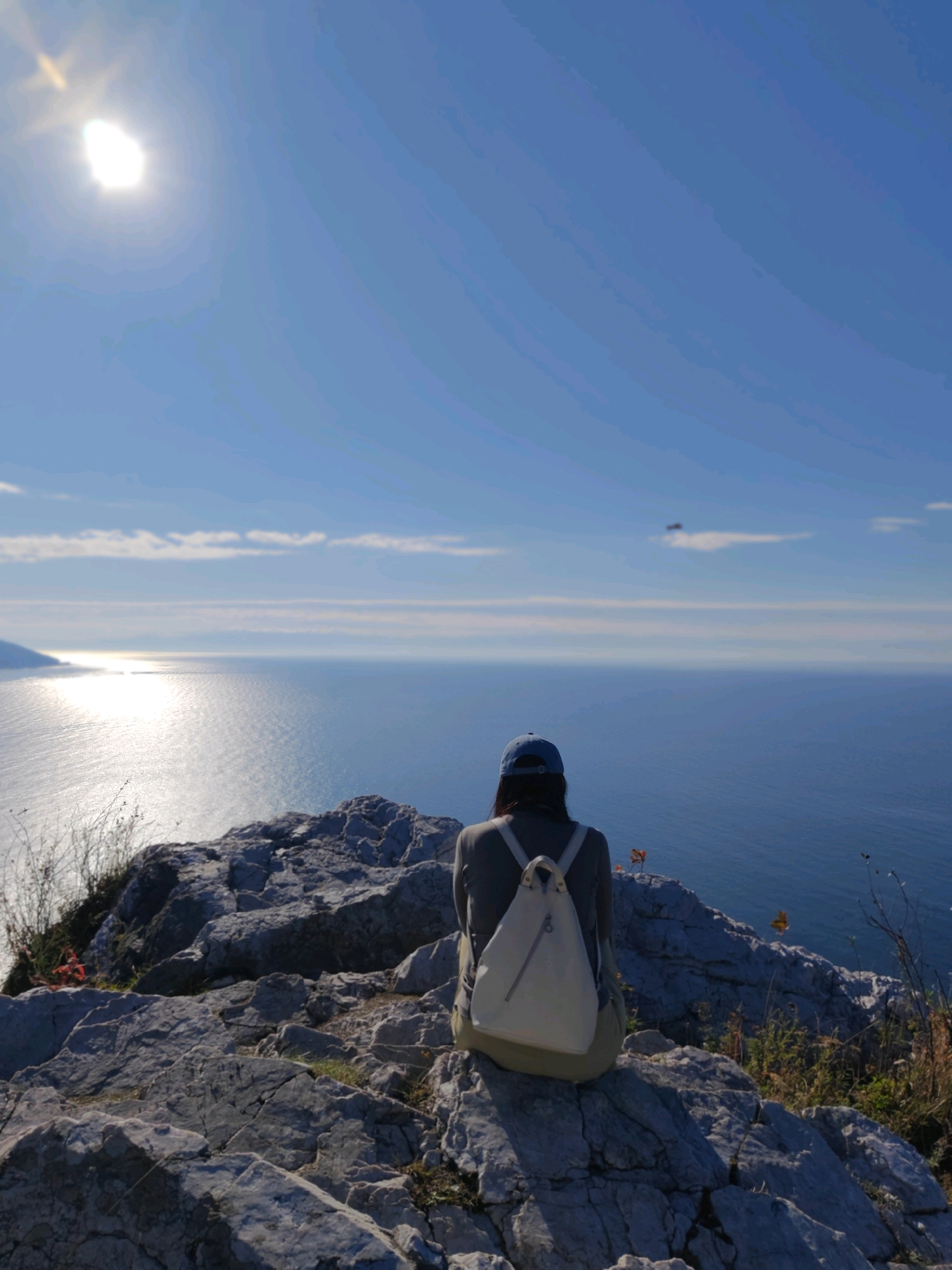A solitary person, viewed from behind, sits on a rocky outcrop overlooking a vast expanse of deep blue water. The individual wears a blue baseball cap and a cream-colored backpack. Sunlight glints off the surface of the water, creating a shimmering path towards the horizon. In the distance, a faint landmass is visible on the left. The sky above is a clear, bright blue with a few wispy clouds and a prominent sun in the upper left quadrant, casting a warm glow. The foreground is dominated by rugged, grey rocks, some sparsely covered with dry vegetation. This scene evokes a sense of peaceful contemplation and immersion in nature, likely a location with a view of Lake Baikal in Russia.
