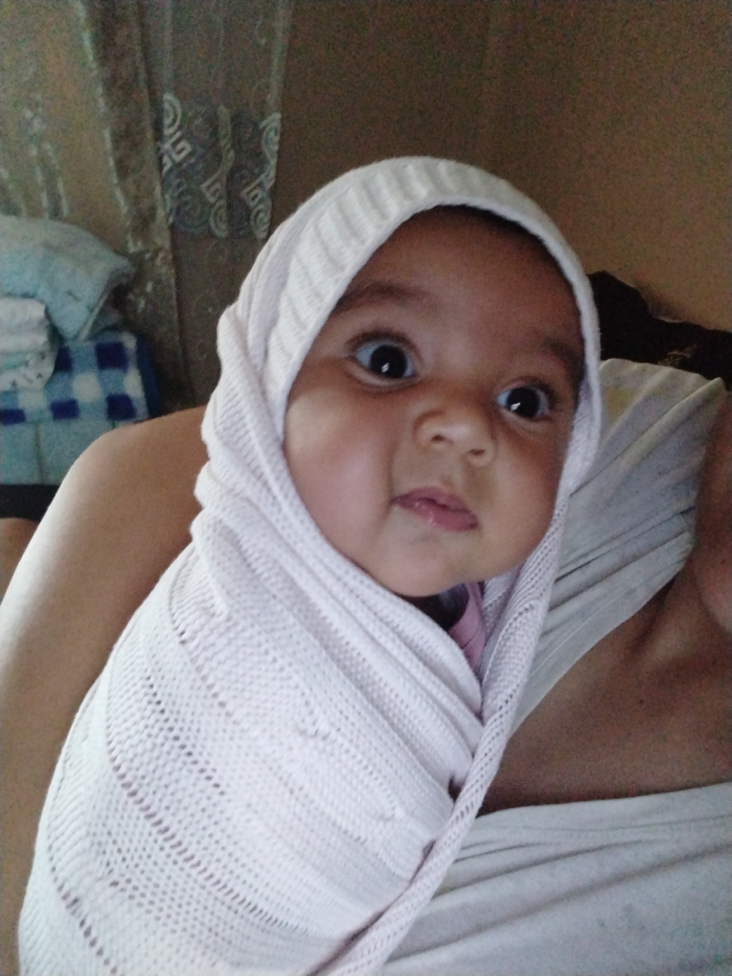 A baby is wrapped in a white knit blanket, covering their head. The baby has wide eyes and a slightly open mouth. The baby is being held by an adult, who is wearing a white shirt. The background includes a curtain and a bed with a blue and white checkered blanket. The location is Unknown, Unknown.