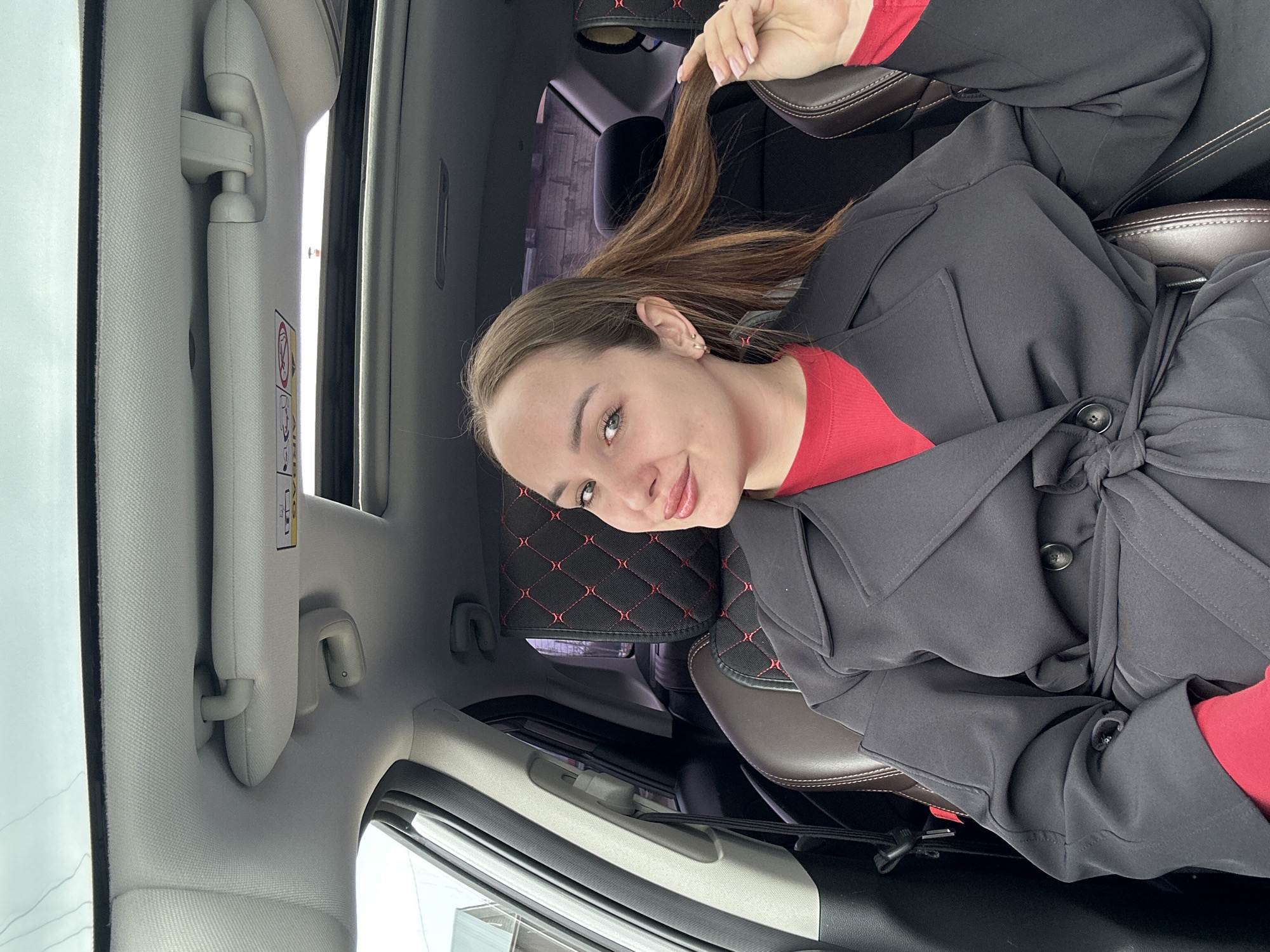 A woman is sitting in the passenger seat of a vehicle in Chaikovsky, Russia. She is wearing a red top and a black trench coat, which is tied at the waist. She is holding a strand of her brown hair. The interior of the vehicle has grey upholstery and brown leather seats with red stitching. The sun visor is down.