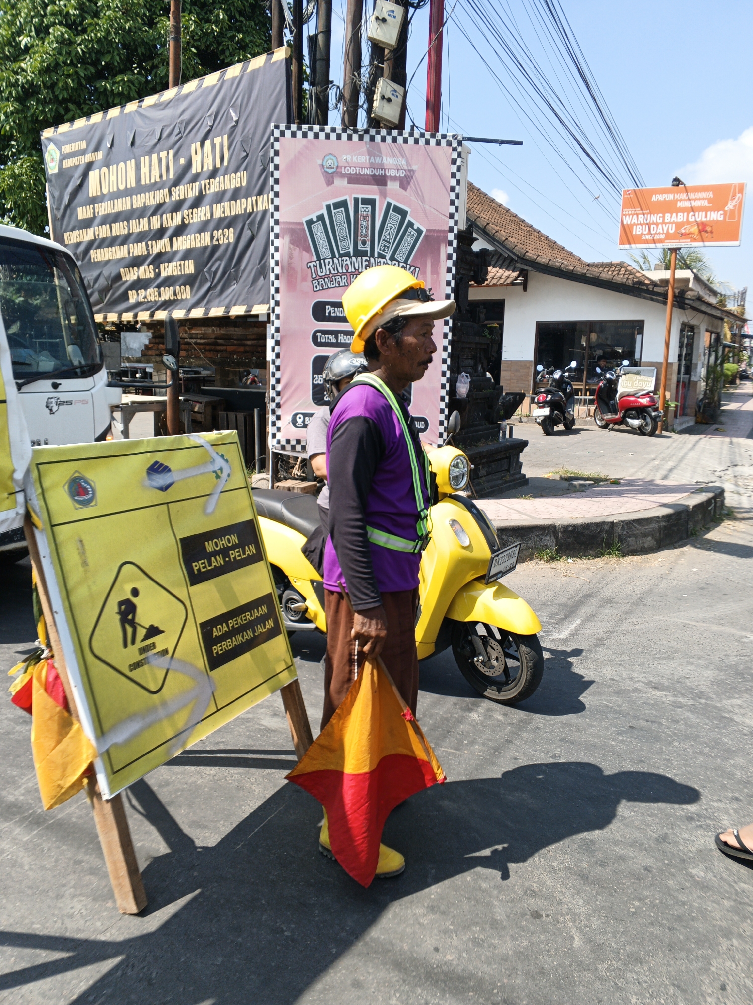An outdoor, street-level, medium shot captures a male construction worker standing next to a yellow scooter on a sunny day. He is wearing a yellow hard hat, a purple shirt, a green reflective vest, and brown pants, with yellow shoes. He holds a red and yellow flag in his right hand. In front of him is a yellow and black sign with a construction icon and text in Indonesian reading "MOHON PELAN-PELAN ADA PEKERJAAN PERBAIKAN JALAN" which translates to "PLEASE GO SLOW THERE IS ROAD REPAIR WORK." To his left, a white truck is partially visible. Behind the worker, a pink and black banner with text and card graphics hangs from a pole. A separate orange sign that reads "WARUNG BABI GULING IBU DAYU SINCE 2000" is also visible. The scene appears to be in the Ubud District of Indonesia, indicated by the presence of local signage and architecture, including a building with traditional Balinese elements. The overall environment is a paved road with some vegetation and utility poles with numerous wires.