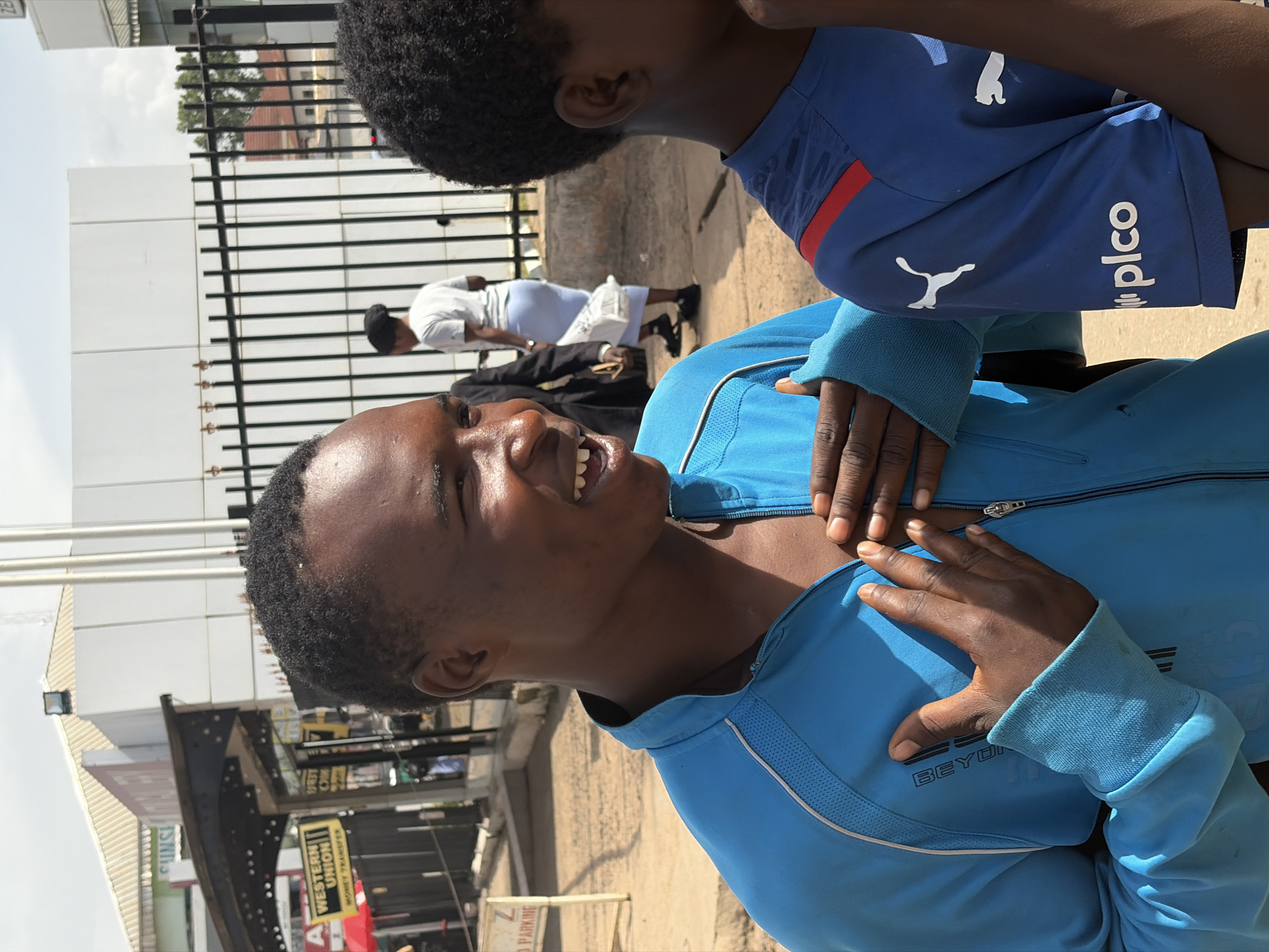 In Jos, Nigeria, a young Black male, wearing a bright blue zipped jacket with white trim, stands with his hands clasped over his chest, smiling broadly. His face is turned to the right, and he has short, dark hair. To his right, a second young Black male is partially visible, wearing a blue t-shirt with red accents and white Puma branding. In the background, several people are walking near a building with a white facade and black bars. A sign for "Western Union Money Transfer" is visible on the left. The ground is paved with concrete and sand.