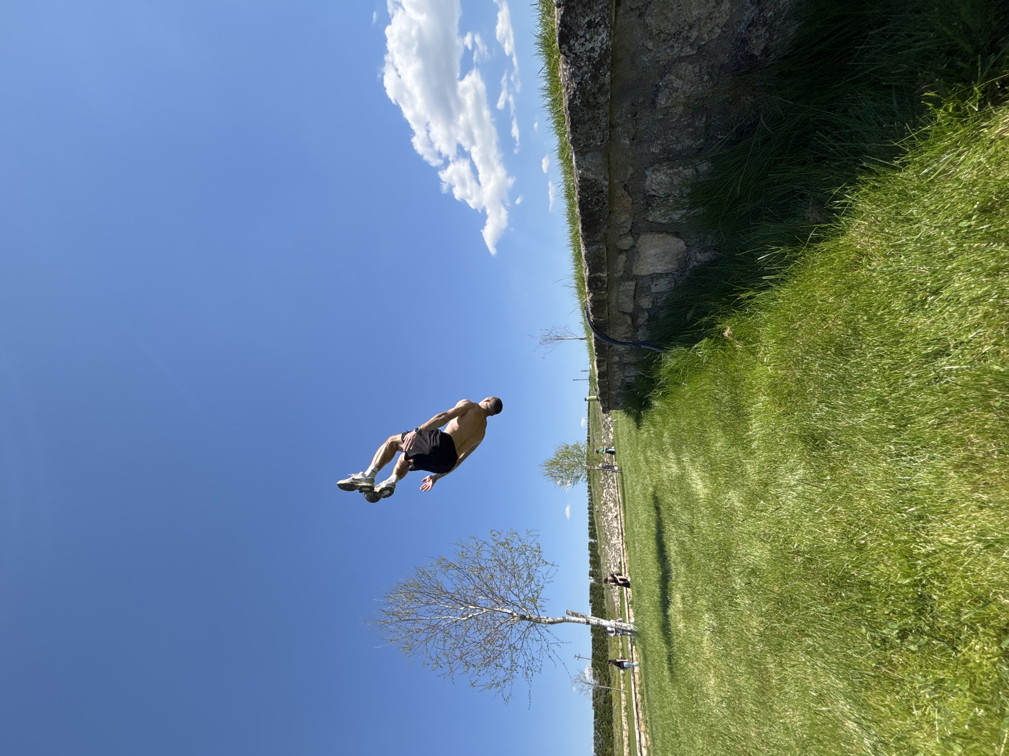 A man is performing a backflip in mid-air in an outdoor setting. He is wearing dark shorts and athletic shoes. The background includes a blue sky with some white clouds, a stone wall with grass growing on top of it, a green grassy area, and several young trees with sparse foliage. There are also other individuals in the background further down the grassy area. The location is identified as being in the city of Unknown, Unknown.