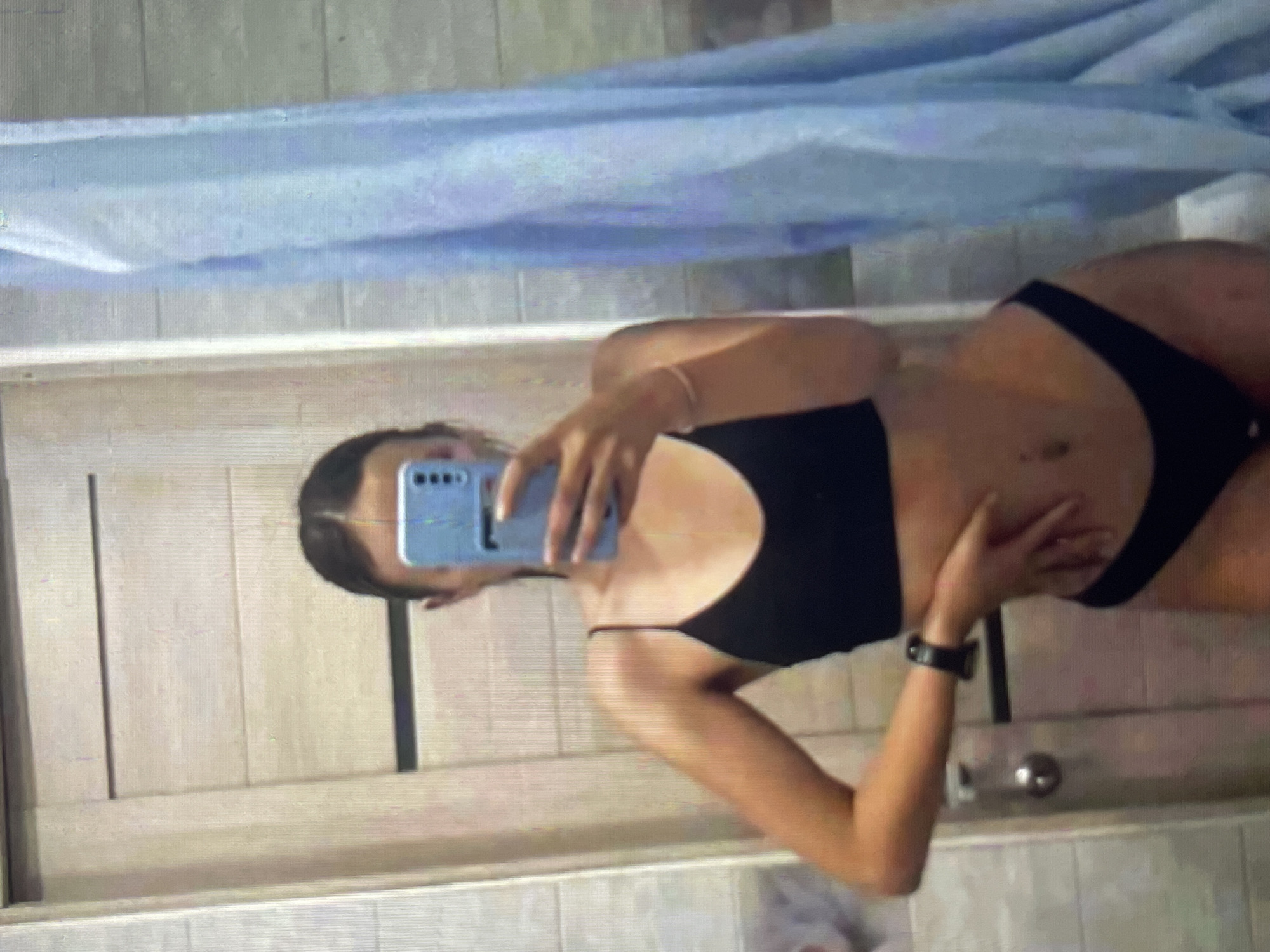 A young woman is taking a mirror selfie in what appears to be a bathroom. She is wearing a black bikini and has a watch on her left wrist. She is holding a light blue smartphone in her right hand, which is obscuring her face. In the background, there is a light-colored door with horizontal panels, a tiled wall, and a light blue shower curtain. The overall lighting is dim, and the image quality is somewhat grainy. There are no identifying landmarks or specific environmental details that would allow for pinpointing the location to Bishkek City, Kyrgyzstan.