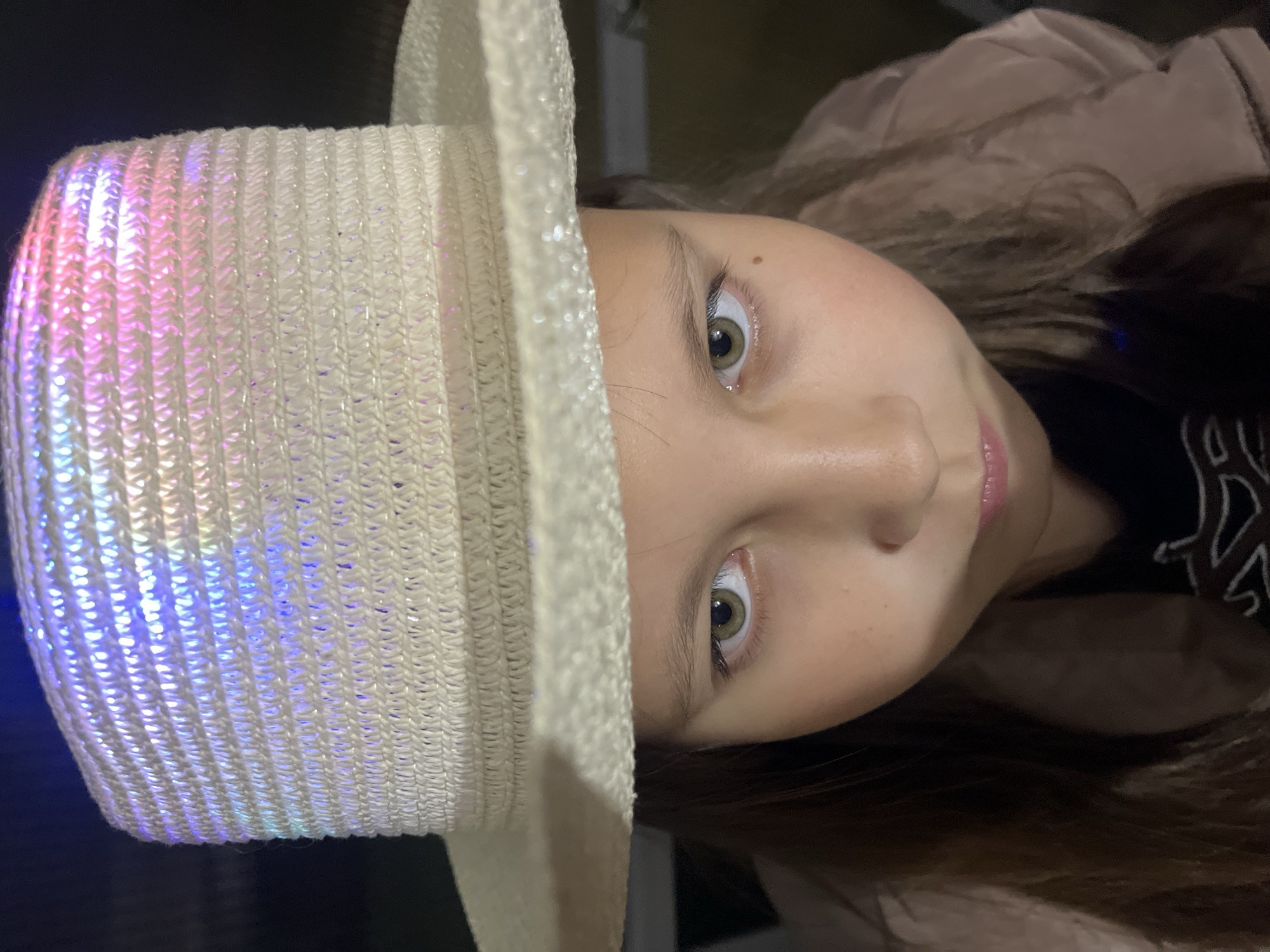 A young girl is the primary subject of the image, positioned in front of a black backdrop. The girl is wearing a tan jacket and has brown hair, with her face the central focus. She is wearing a white straw hat with blue and purple lights in the crown. The image is taken at an angle, with the subject's head tilted slightly to the side. The location is not provided in the image.