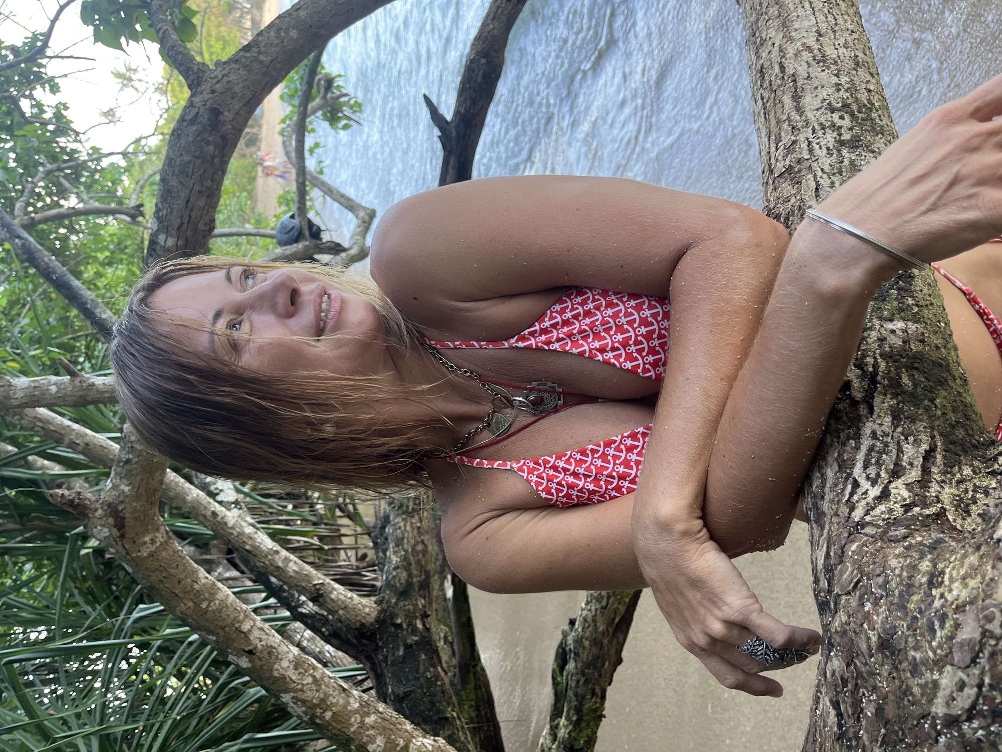 A woman with blonde hair and braces is leaning on a fallen tree trunk on a sandy beach, with the ocean in the background. She is wearing a red and white anchor-patterned bikini and several necklaces. Her arms are crossed, and she is wearing a ring on her left hand and a bracelet on her right wrist. The water is calm and slightly wavy, and there are people visible in the distance on the beach. The scene appears to be set in Goviyapana, Sri Lanka, as suggested by the presence of tropical foliage.