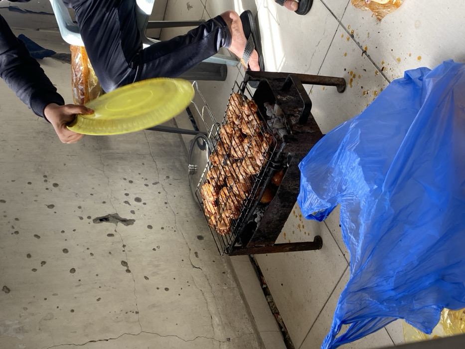 The image depicts an outdoor barbecue scene in what is identified as Khan Yunis, Palestinian Territory. In the foreground, a rusted, rectangular metal charcoal grill with four legs holds a metal grate laden with numerous pieces of cooking, browned chicken, possibly wings or drumsticks. Below the chicken, burning charcoal and several round vegetables are visible. A silver-colored pot lid rests on the side of the grill.
A partially visible individual, seated on a light blue plastic chair, wears dark long-sleeved clothing, dark pants, and black flip-flops. This individual's right hand actively wields a bright yellow, oval-shaped plastic plate, blurred due to motion, presumably used for fanning the coals. A second pair of feet in black flip-flops is partially visible on the right side of the frame, suggesting at least one additional person nearby.
The ground consists of light-colored tiles, exhibiting numerous dark splatters, likely grease or drippings, particularly around the barbecue. A large, crumpled bright blue plastic bag occupies the immediate foreground. In the background, a rough, light grey concrete wall is visible, showing numerous small, irregular dark marks and several distinct cracks, including a more substantial, irregular hole. Strong daylight illuminates the scene, casting discernible shadows.
