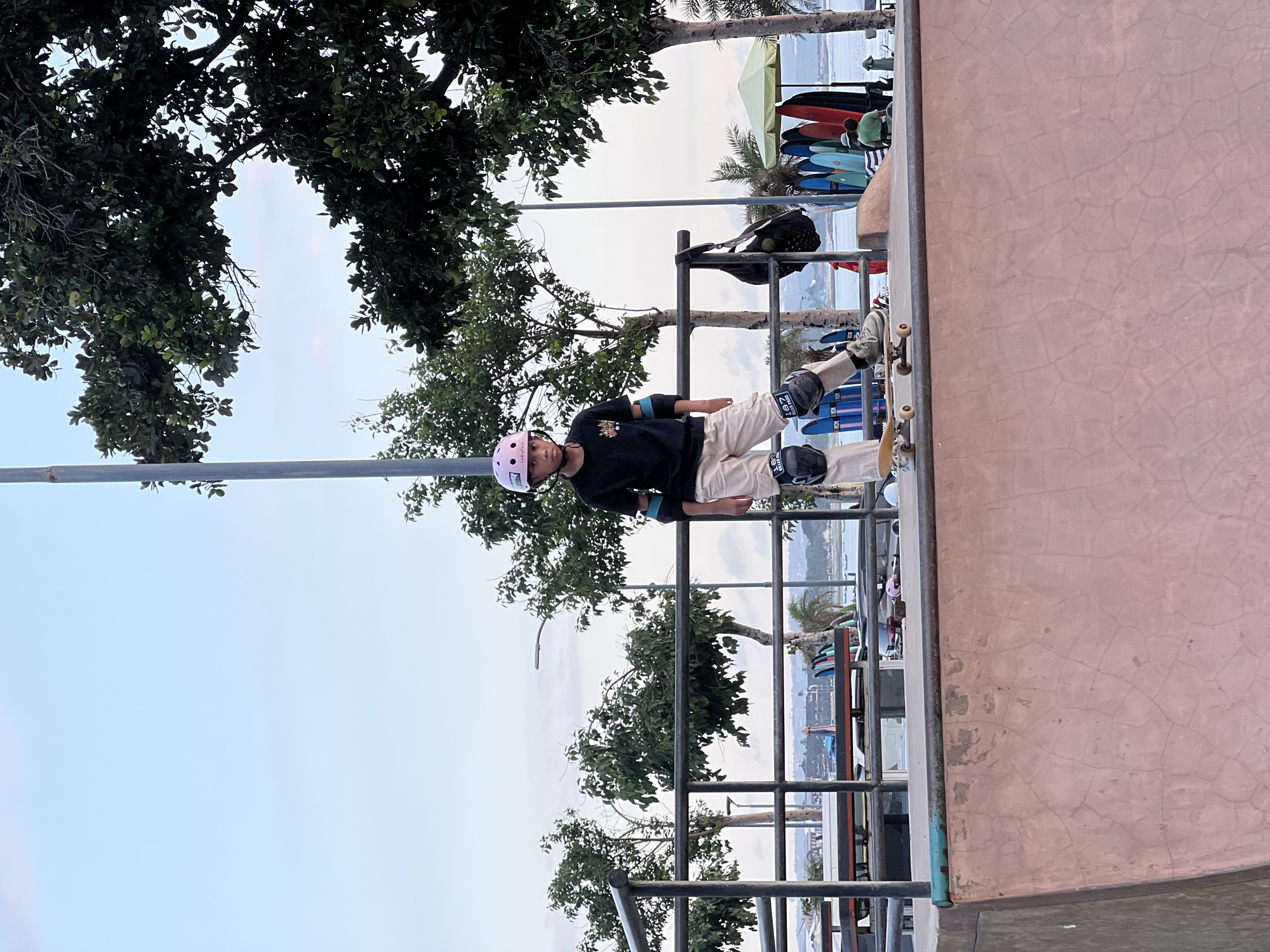 A young person wearing a pink helmet, black long-sleeved shirt, beige pants, and protective gear on the knees and elbows stands on a skateboard at a skatepark. They are positioned at the edge of a ramp with metal railings. In the background, there is a beach scene with palm trees, water, and several surfboards leaning against a structure. The location appears to be Kuta, Indonesia, due to the coastal environment and surfboards, consistent with the surf culture of Kuta. There is an overhanging tree in the background.